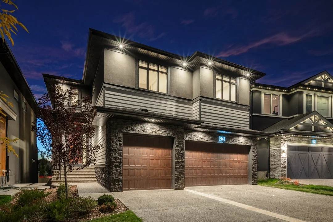 Exterior view of a modern two-story house at dusk with illuminated lights, two brown garage doors, and a small front yard with a tree and plants.
