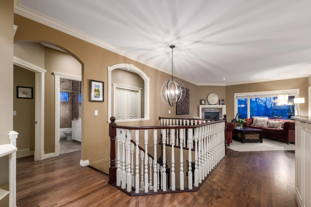 Living room with beige walls, wooden staircase with white and dark wood railing, ceiling light fixture, fireplace, windows, and a red sofa with decorative pillows.
