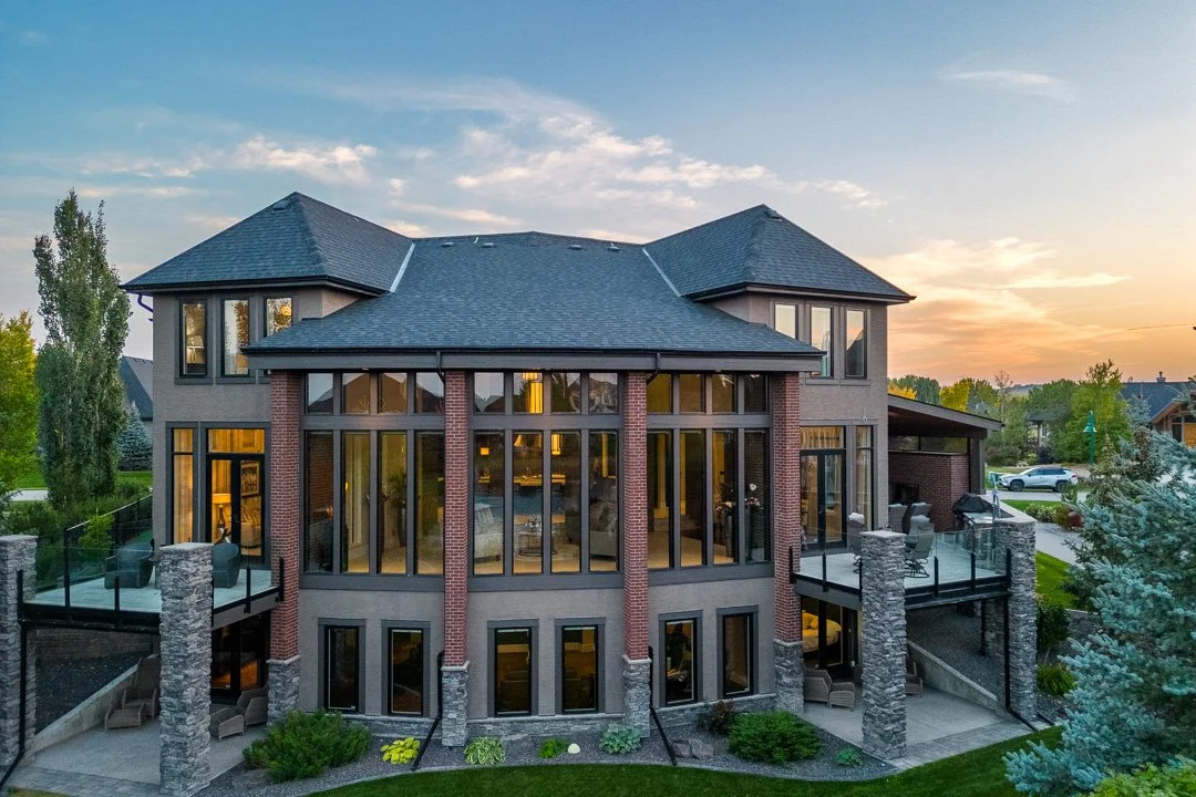 Large modern house with stone and brick exterior, multiple large windows, and a spacious balcony with glass railing, set in a suburban neighborhood at sunset.