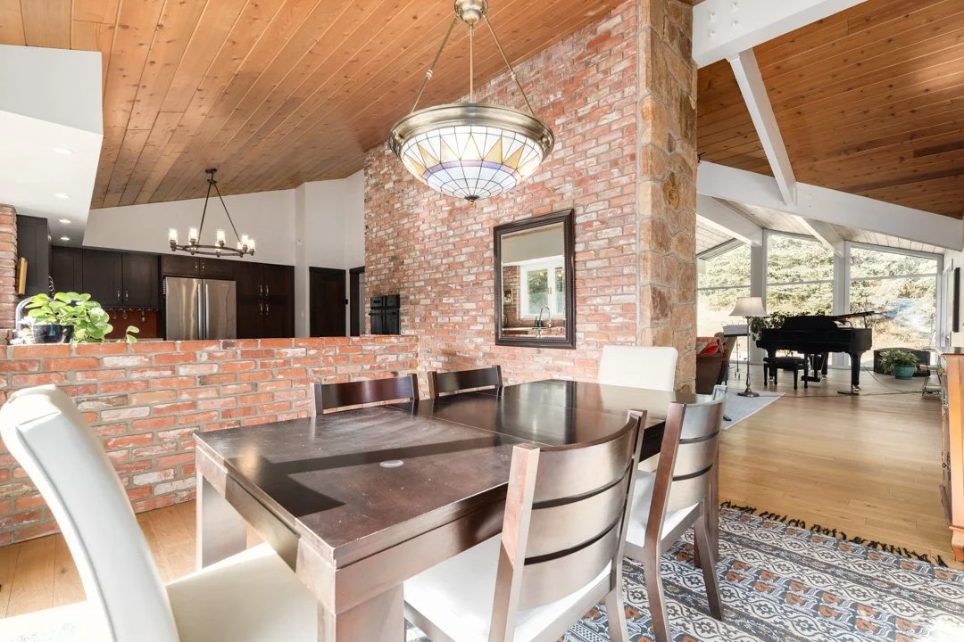 Dining room with a dark wood table, white and dark wood chairs, brick walls, wooden ceiling, and a chandelier. Visible kitchen area with dark cabinetry and stainless steel refrigerator in the background.