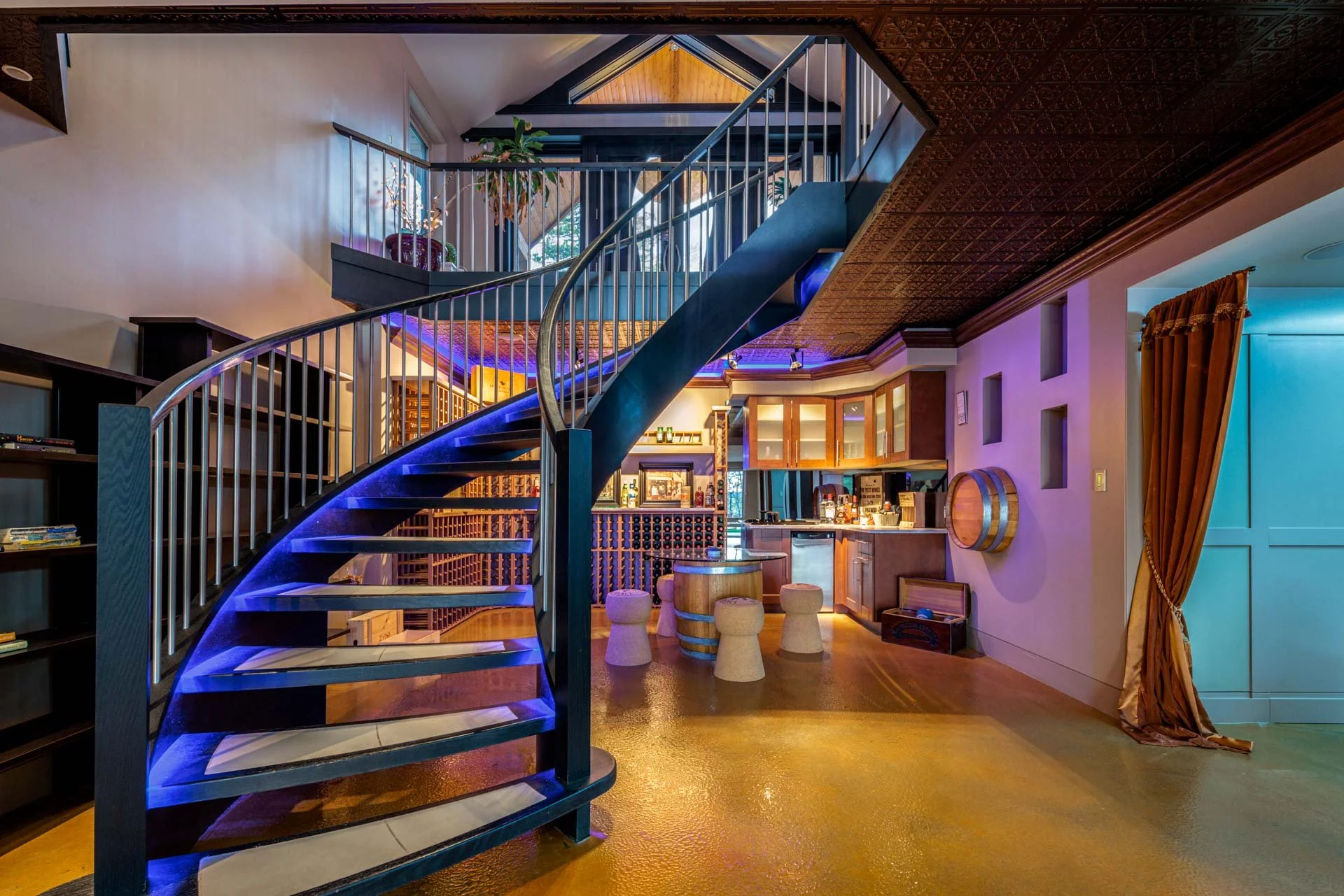 Interior view of a multi-level home with a black spiral staircase with blue LED lighting, a kitchen area with wooden cabinets, a bar with stools, and decorative wine storage with bottles and books.