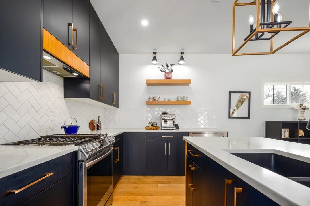 Modern kitchen with black cabinets, white countertops, and wooden accents. Open shelving with cups and decor, stainless steel appliances, and a window with a view of trees.