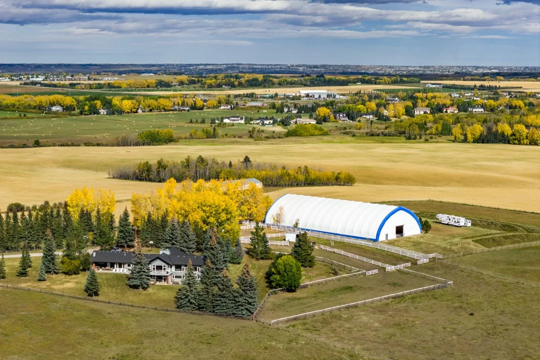 Aerial view of a rural area featuring a stable with a white and blue roof, a house surrounded by trees, and expansive fields with some trees turning yellow, indicating fall. The landscape stretches to the horizon under cloudy skies.