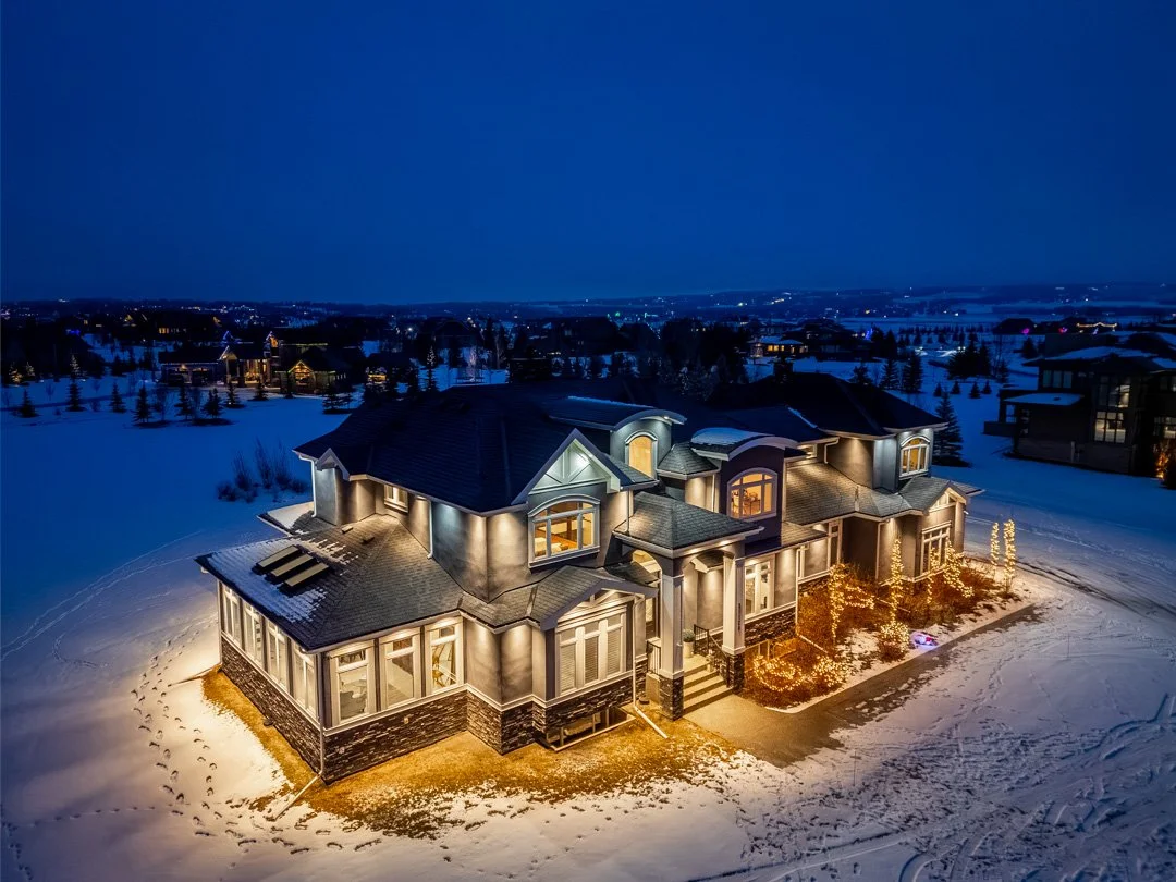 A large, illuminated house in a snowy landscape at night with multiple windows and a dark roof, surrounded by snow-covered ground and trees.