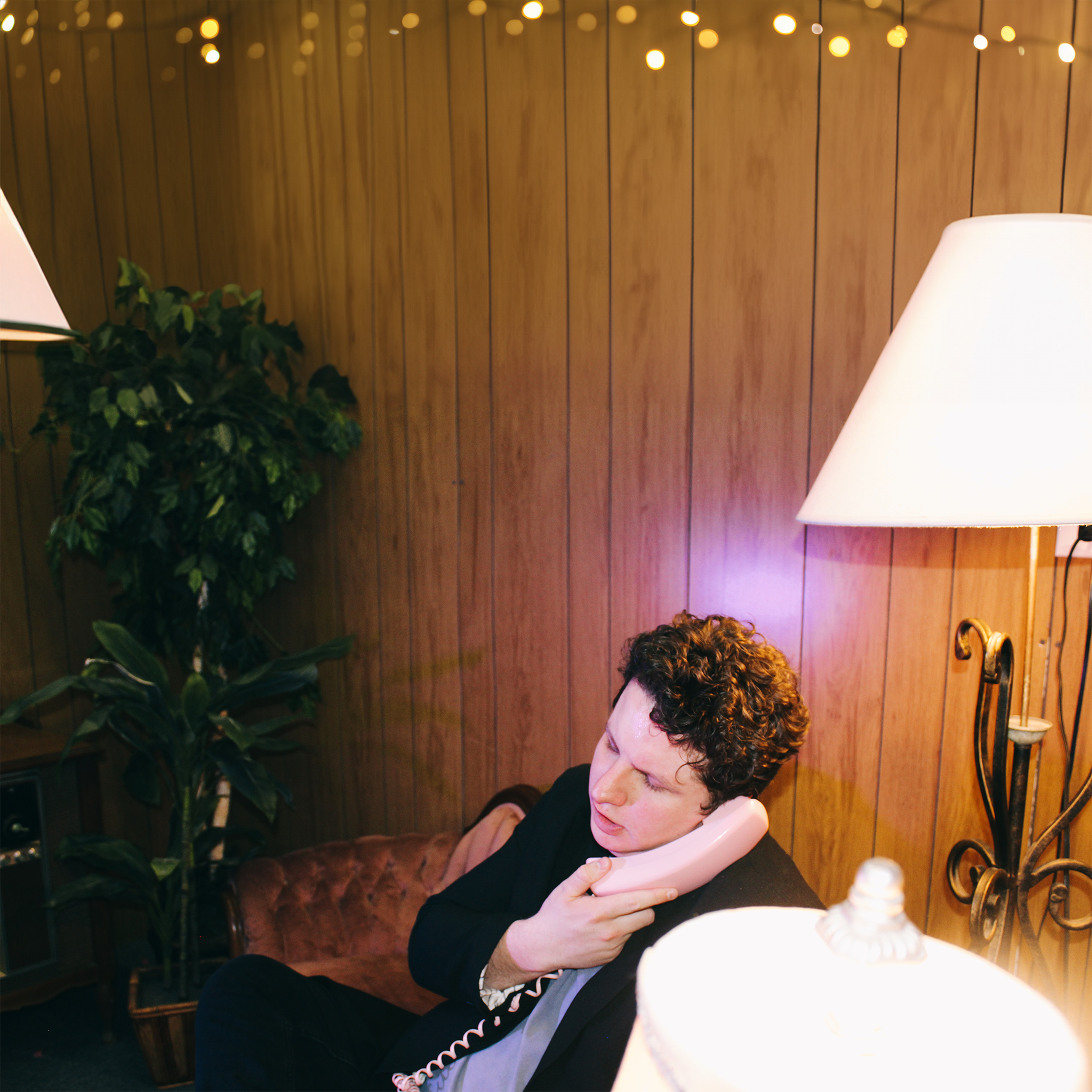 A young man Griffin Oswald, with curly hair sitting on a brown sofa, talking on a pink rotary telephone in a room with wood-paneled walls, a tall potted plant, and two table lamps, one on each side of him. String lights are hanging from the ceiling.