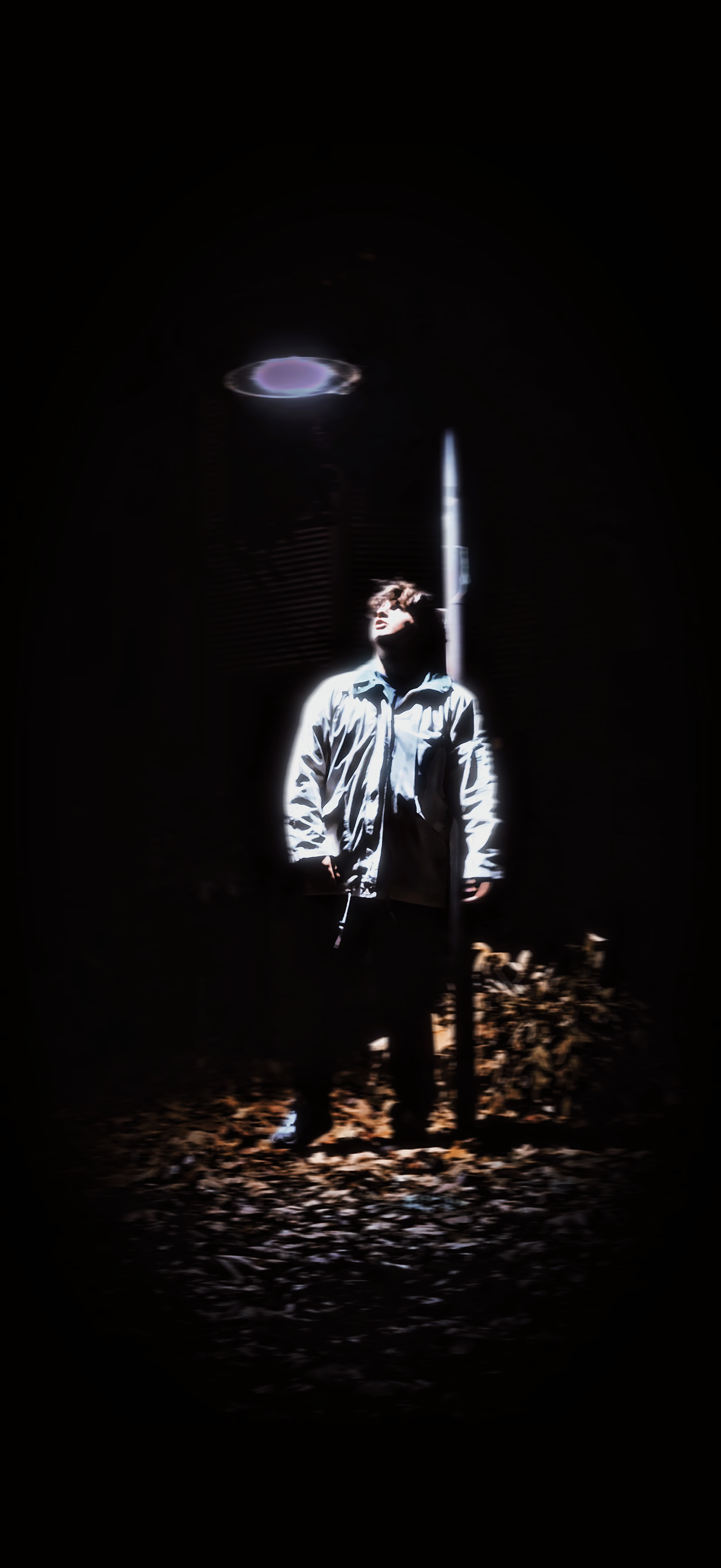 A person Griffin Oswald stands outdoors at night, illuminated by a streetlamp, with fallen leaves on the ground and a dark sky above.