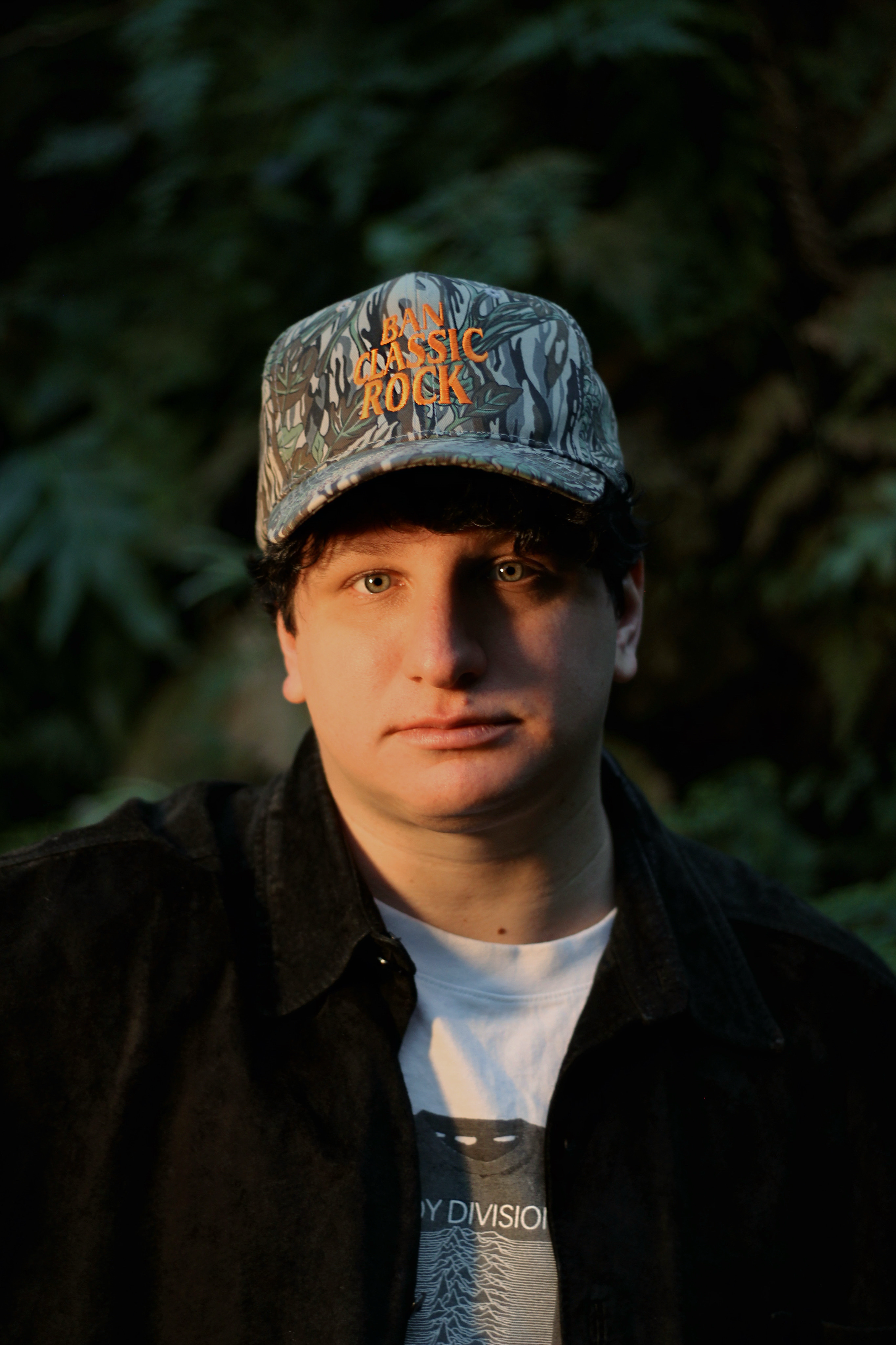 A young man, Griffin Oswald, with light skin and light-colored eyes wearing a patterned cap that says "BAN CLASSIC ROCK" and a black jacket over a white t-shirt, standing outdoors with a blurred nature background.