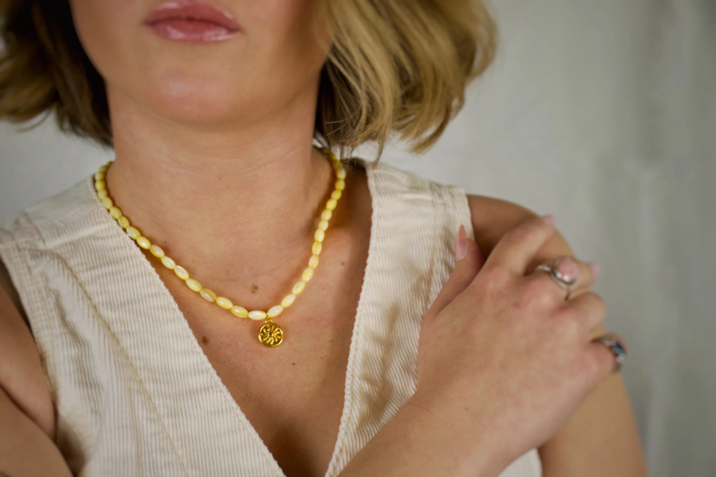 A woman wearing a yellow beaded necklace with a round gold pendant and a sleeveless beige top. She is touching her shoulder with her right hand and has rings on her fingers.