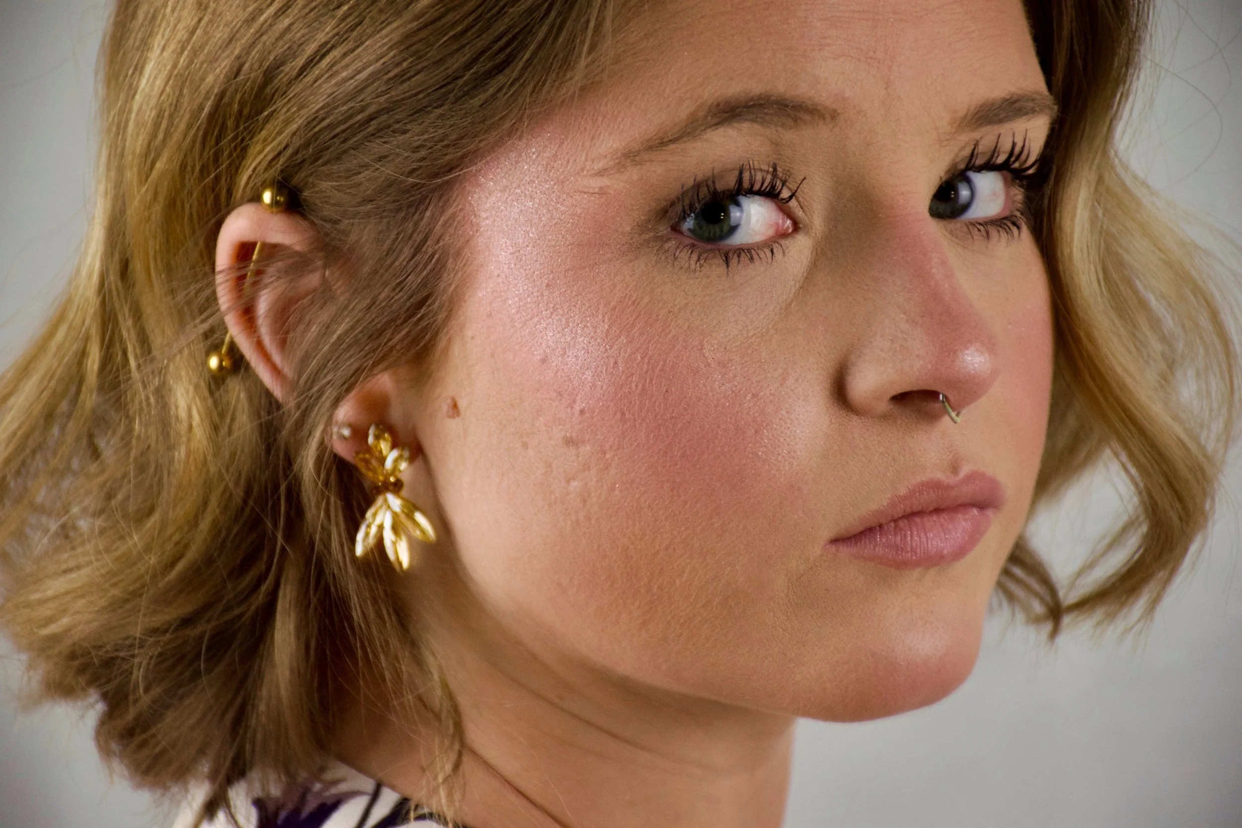 Close-up of a woman with light skin, short wavy hair, and multiple earrings, including gold floral earrings and gold ear cuffs, wearing mascara and natural makeup.