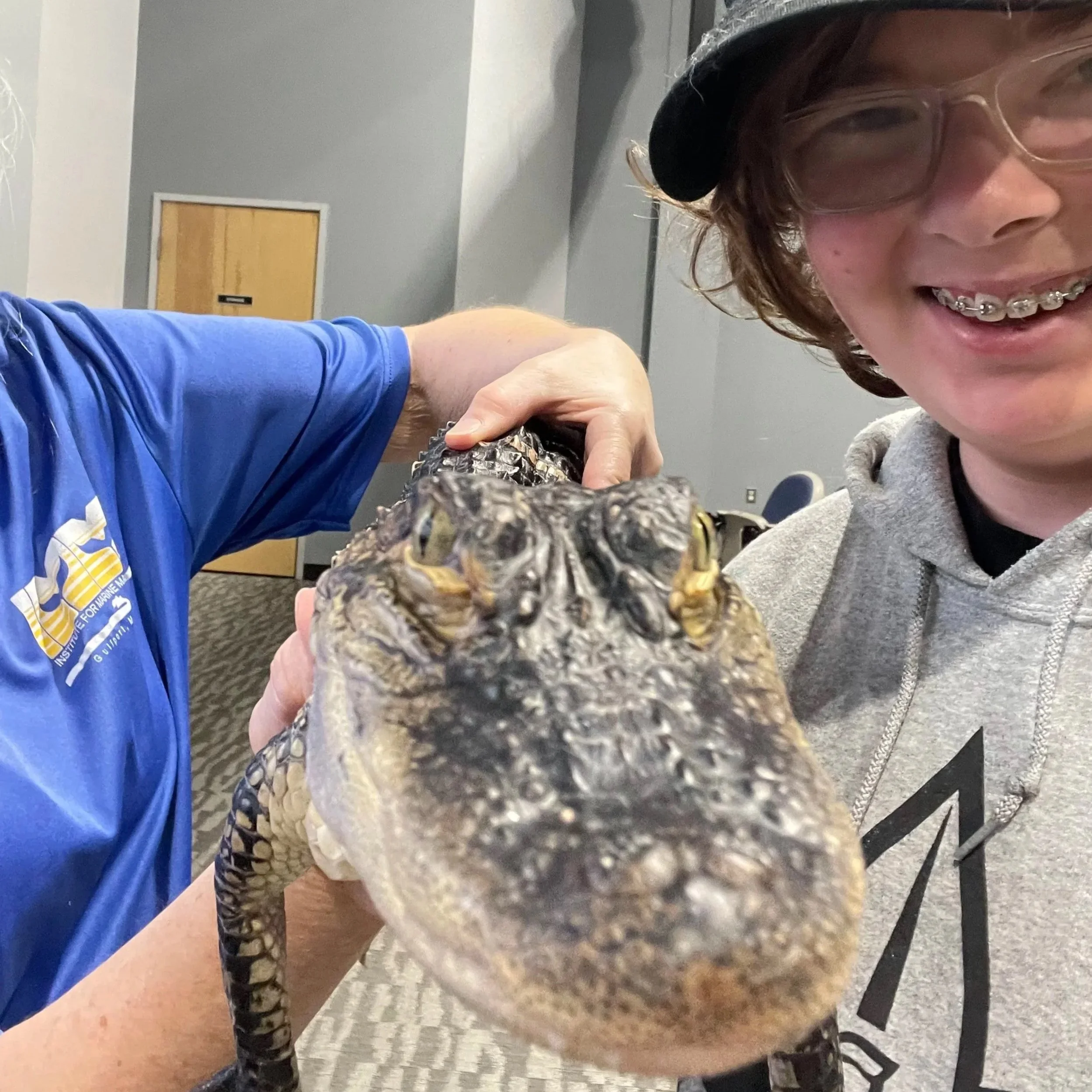 Close-up of a young person with braces smiling, wearing glasses, a black beanie, and a gray hoodie, holding a snake in their hand inside a room with gray walls, a door, and a patterned carpet.