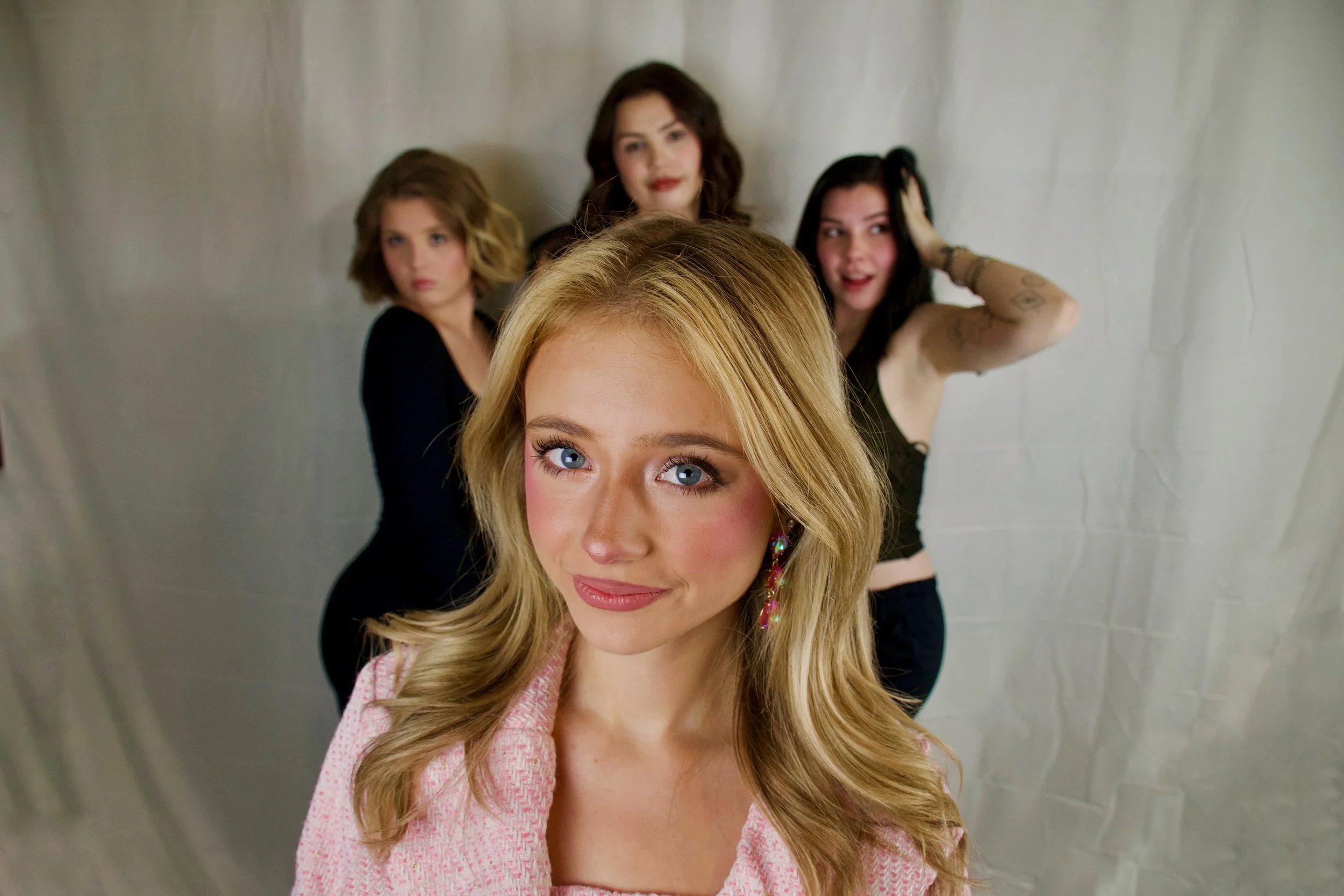 A young blonde woman with blue eyes and pink lipstick smiling at the camera, standing in front of four women with varying hair colors and styles, all posing in front of a plain backdrop.