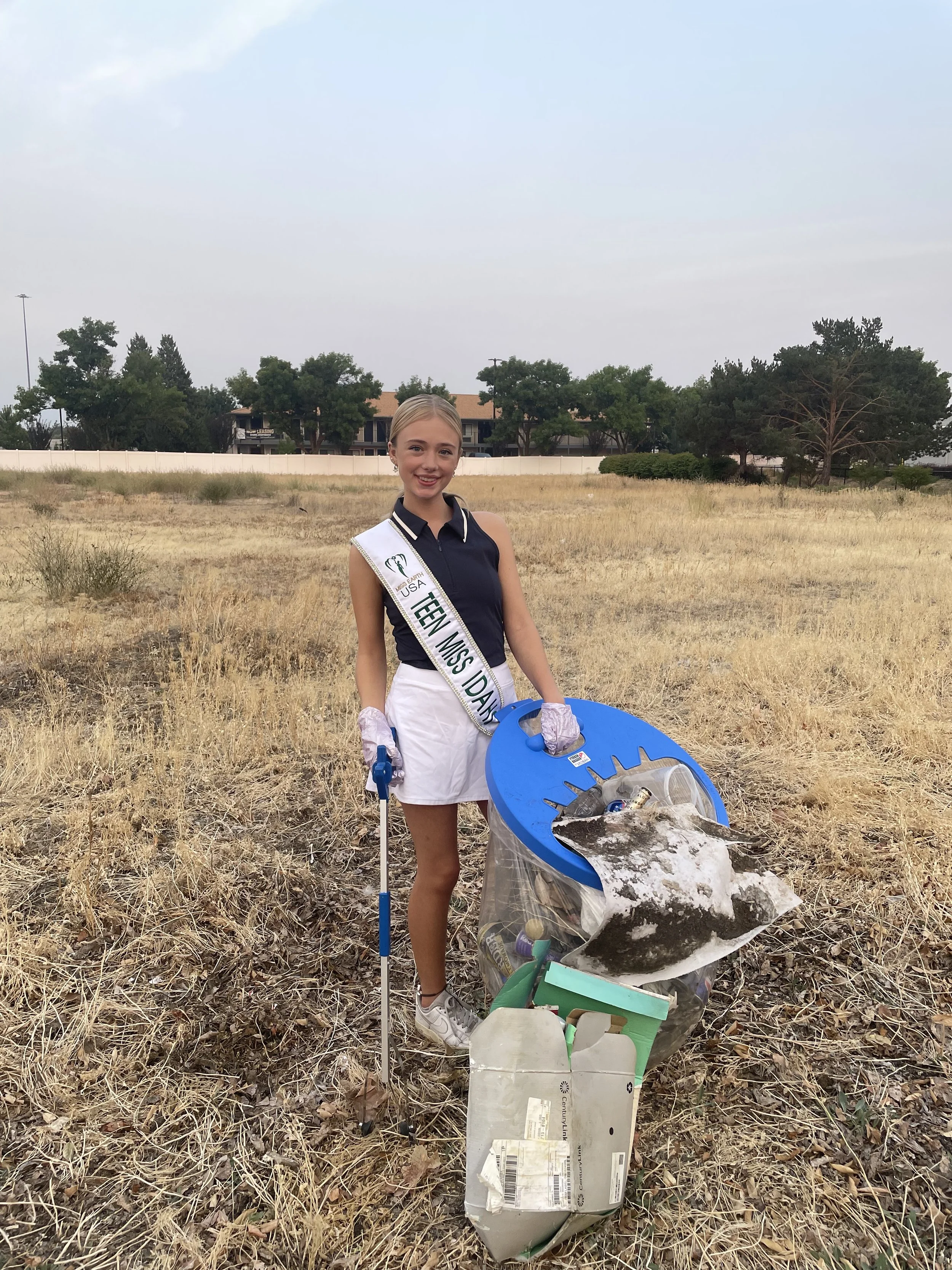 Young woman wearing a sash that reads "Teen Miss Davi" and gloves, standing in a field with a trash grabber, collecting litter, next to a blue trash container filled with trash.