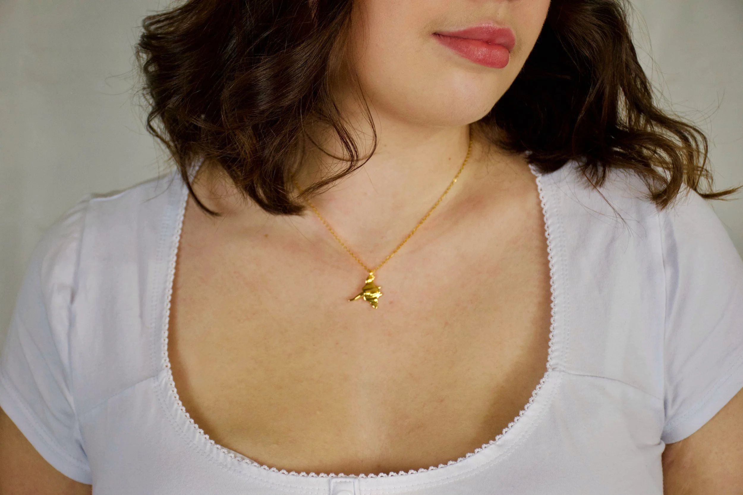Close-up of a woman wearing a white top with a square neckline, gold necklace with a hummingbird pendant, and with wavy brunette hair.