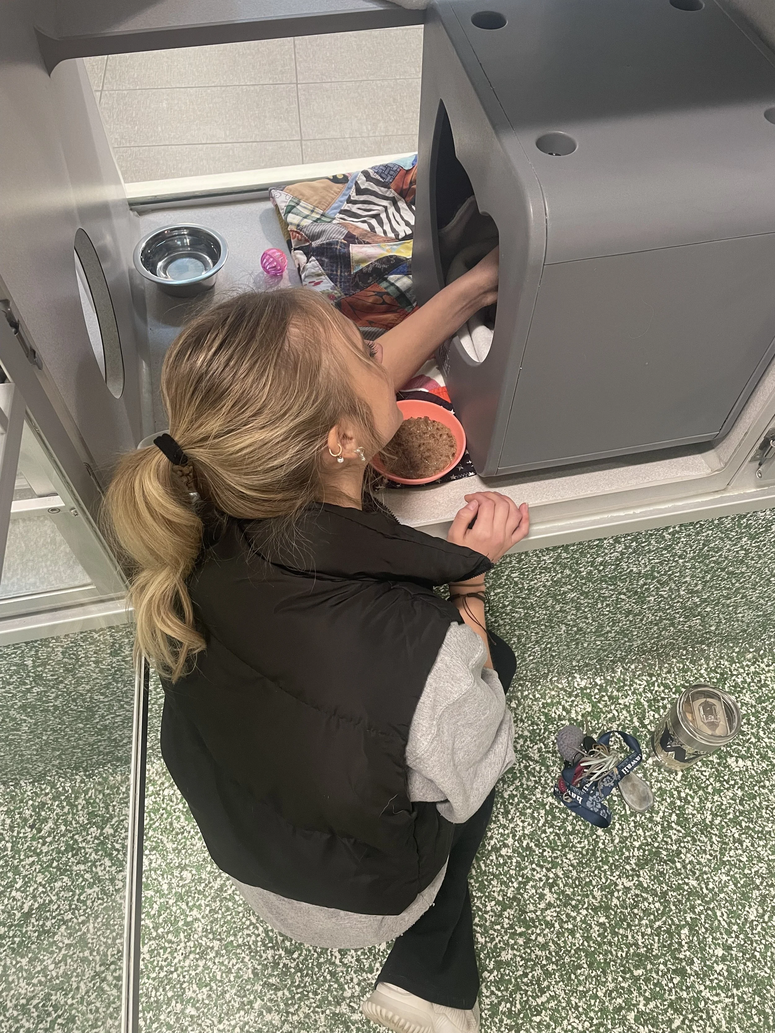 A woman is lying on the floor next to a dryer, reaching inside with her arm. She has a ponytail, wears a black vest over a gray hoodie, and appears to be eating from a bowl of food. Items nearby include a water bottle, keys, a travel mug, and a pair of sunglasses.