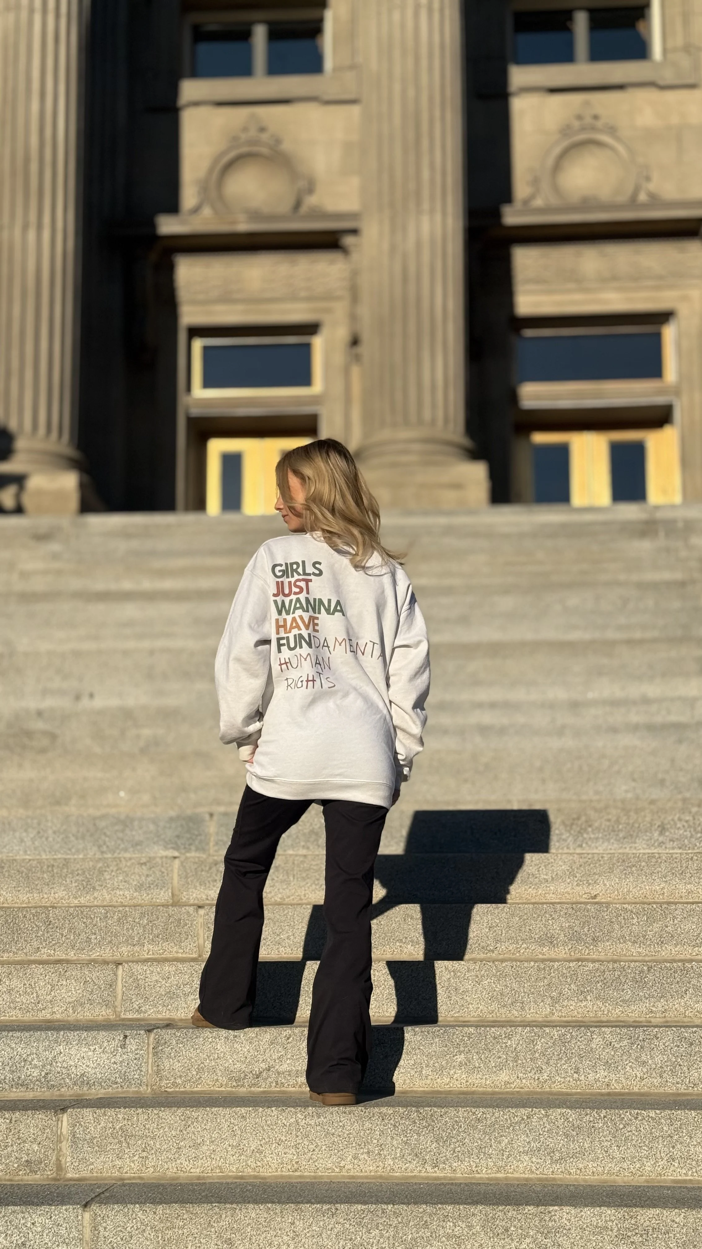 A woman with blonde hair stands on the steps of a historic building with classical columns, wearing a gray sweatshirt with the phrase "Girls Just Wanna Have Fundamental Human Rights" written on the back. The sunlight casts a shadow of her on the steps.