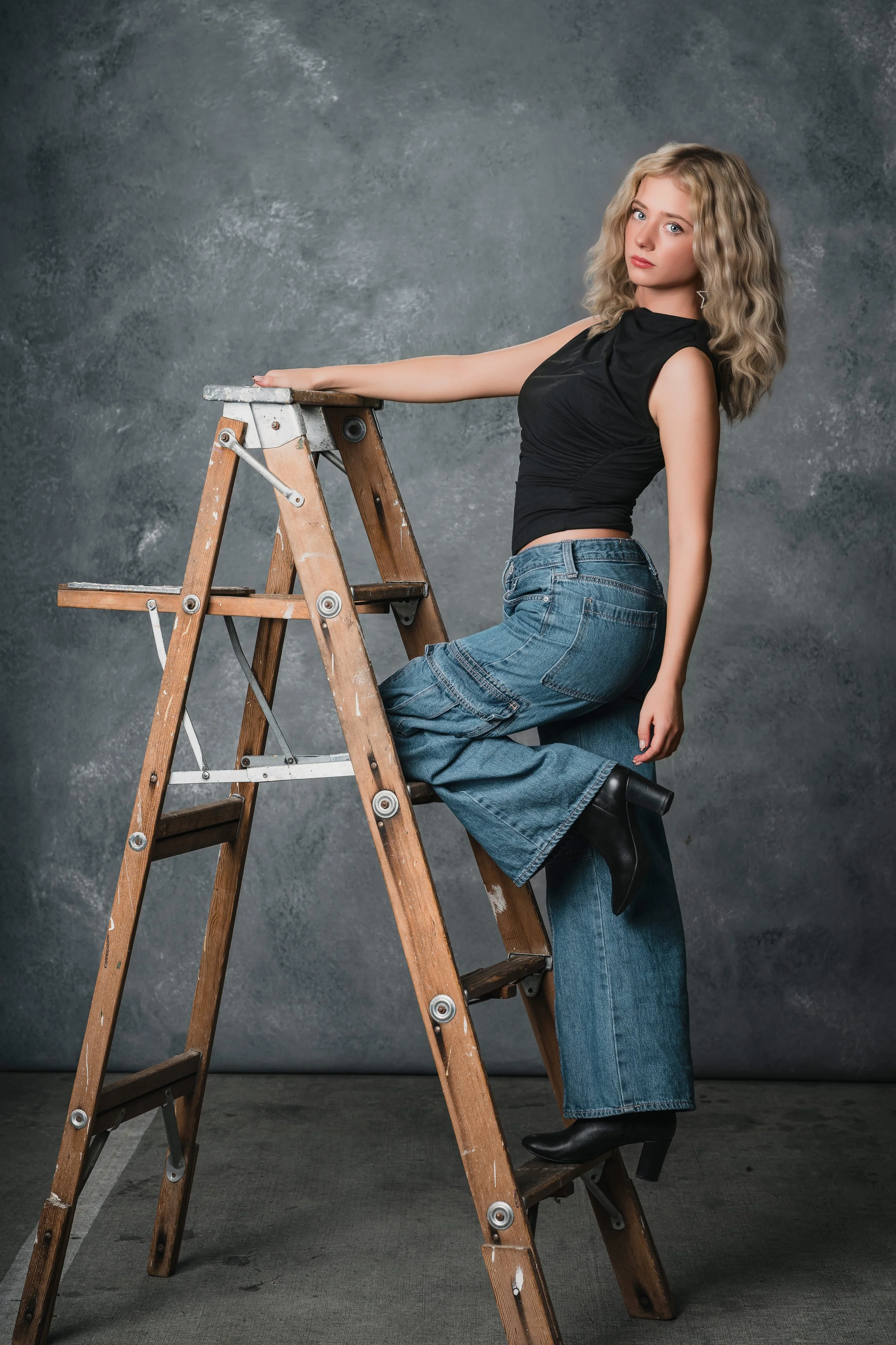 A young woman with curly blonde hair wearing a black sleeveless top, wide-leg jeans, and black heeled boots, sitting on a wooden step ladder against a textured gray background.