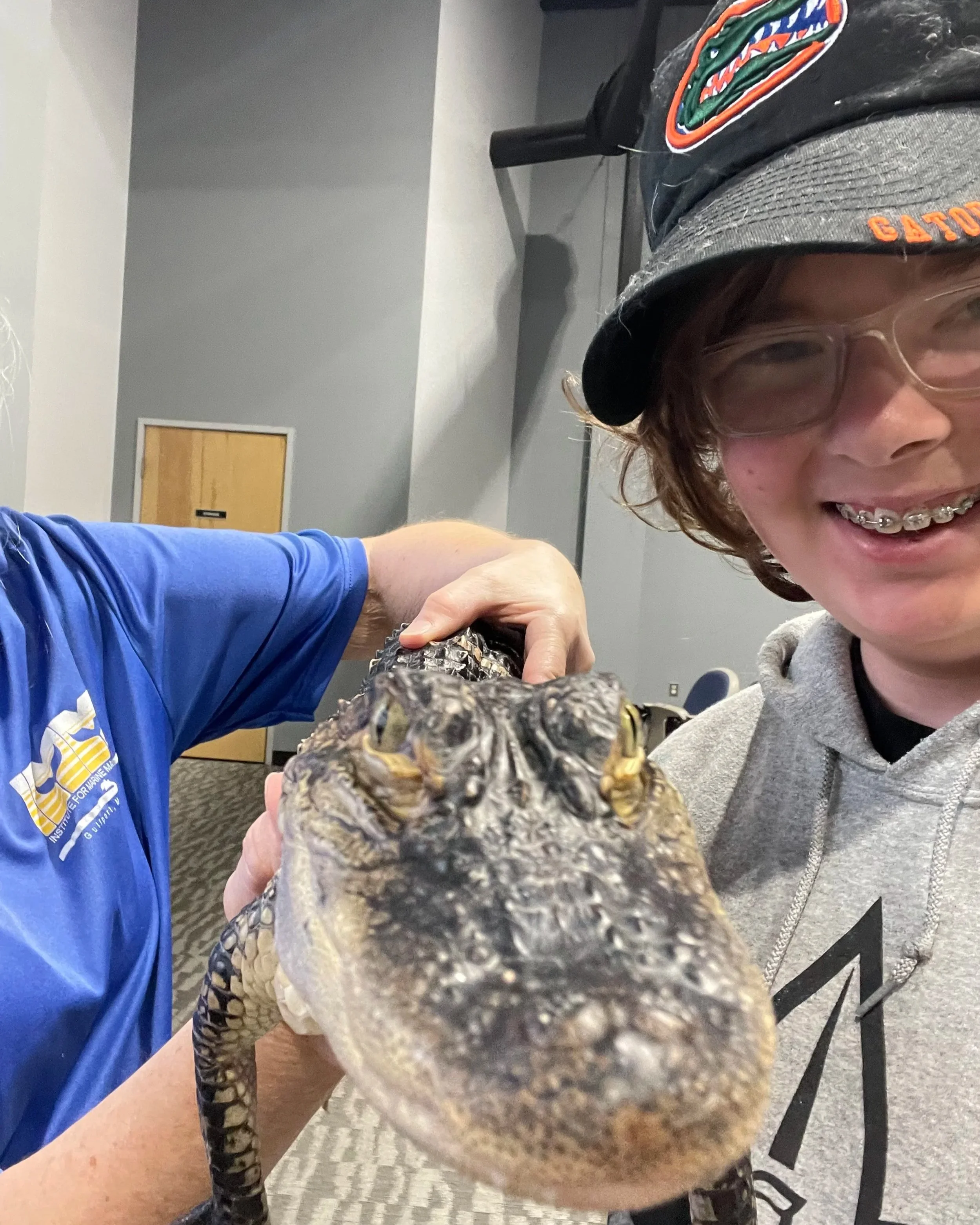 A person taking a selfie with a young crocodile in an indoor setting. The person is smiling, wearing glasses, a gray hoodie, and a black cap with an orange and green logo. The crocodile's head is close to the camera, with its eyes and snout visible.