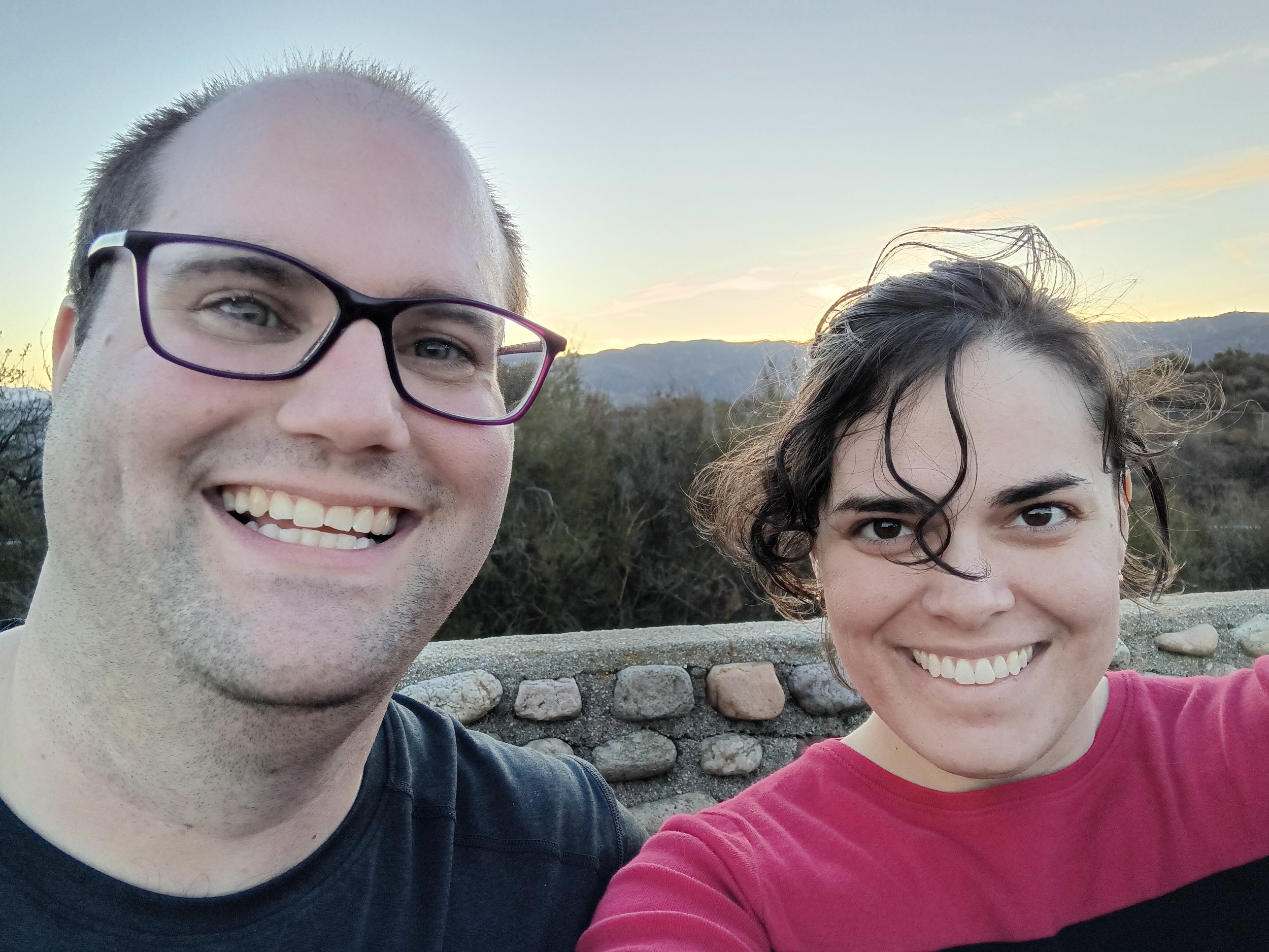 The image shows a woman with brown wavy hair standing in front of a sunset
