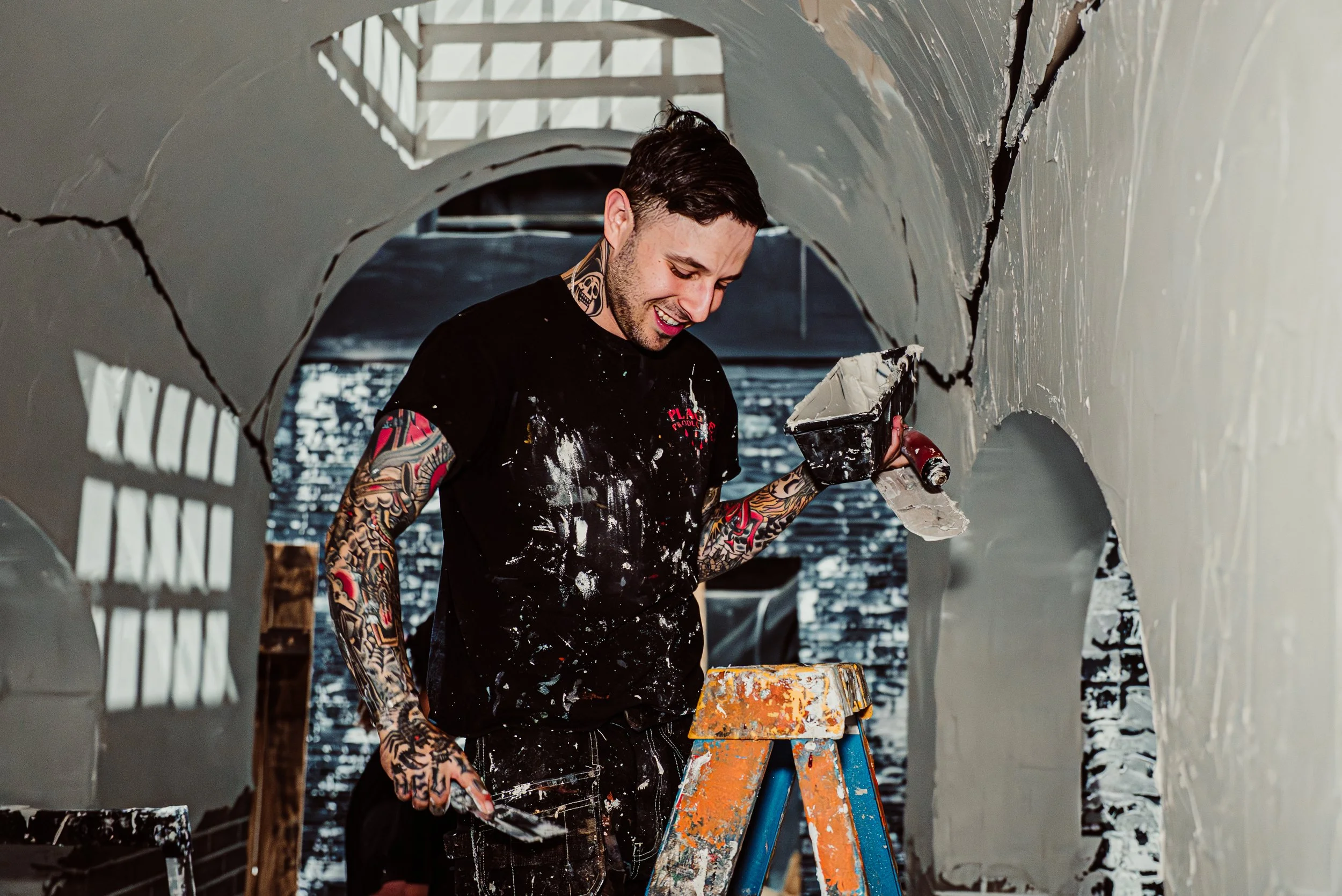 A man with tattoos on his arms and neck working on a wall, smiling, holding a plastering trowel and a bucket of plaster, standing on an orange and blue ladder in a room with arched doorways.