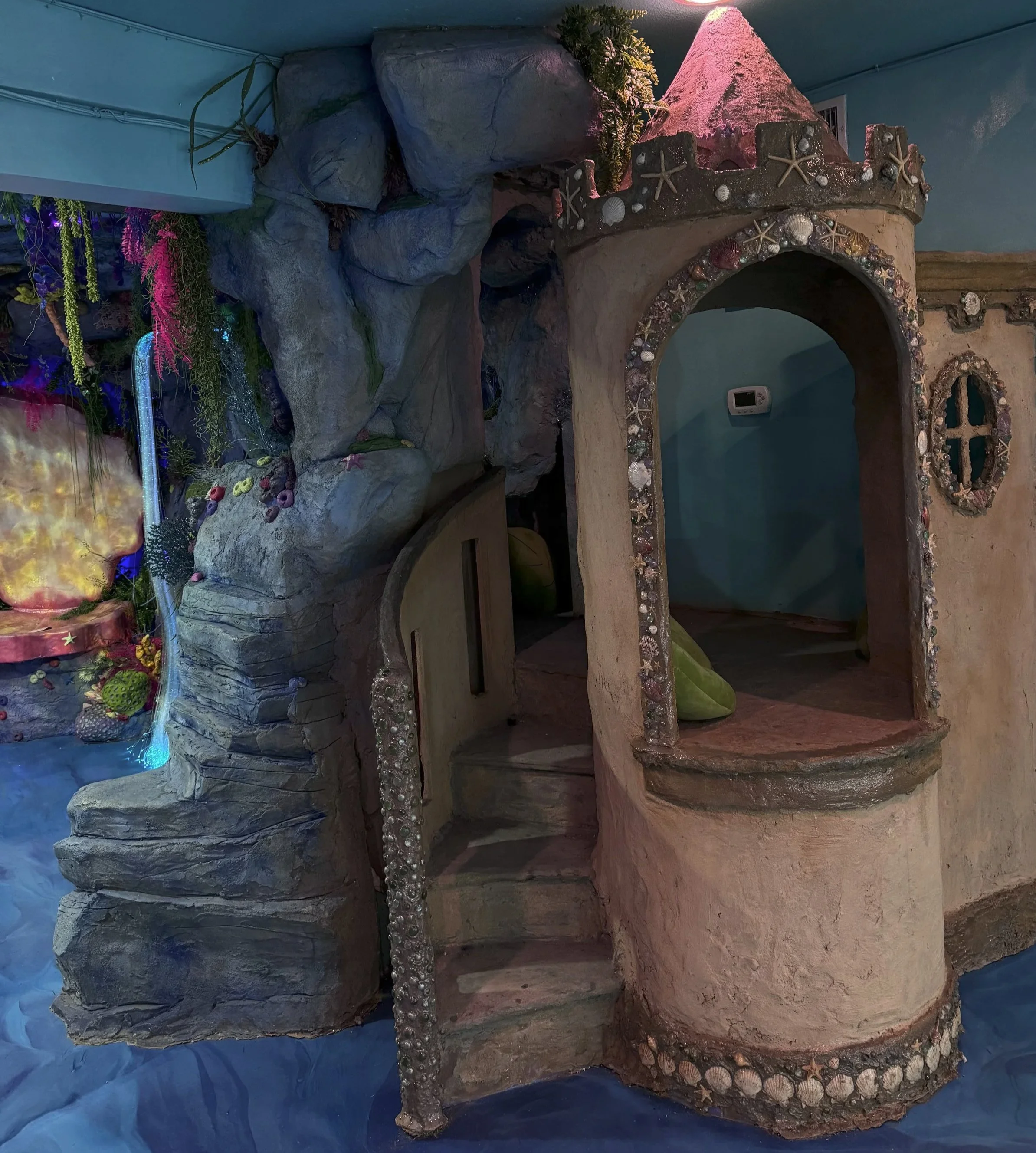 A whimsical fantasy-themed indoor playhouse designed to look like a fairy tale castle with stone and shell accents, set against a rocky backdrop with colorful plants and decorations.