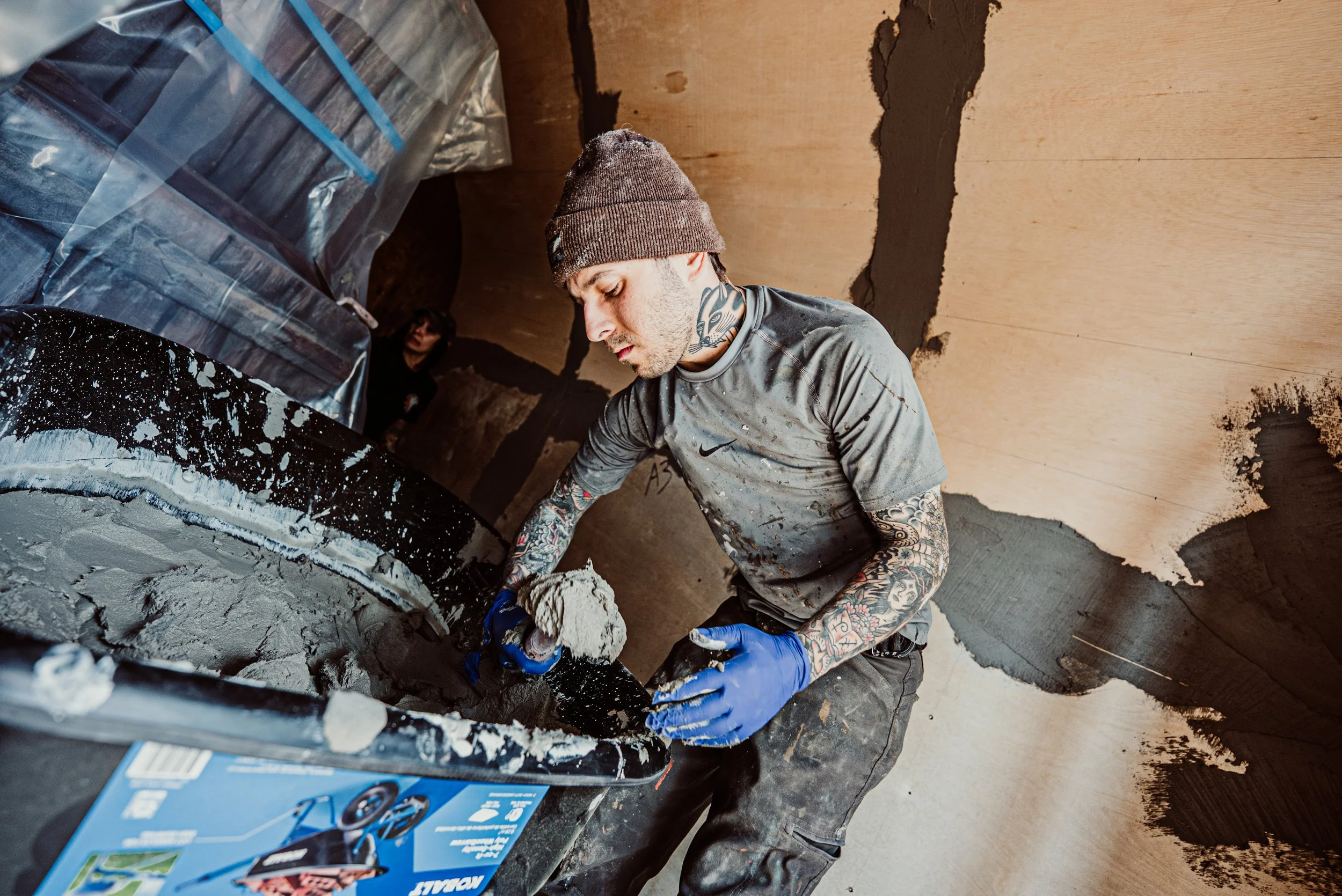 A man with tattoos, wearing a brown beanie, gray distressed shirt, and blue gloves, working with cement or plaster in a construction or renovation site with plywood walls and stacked boxes nearby.