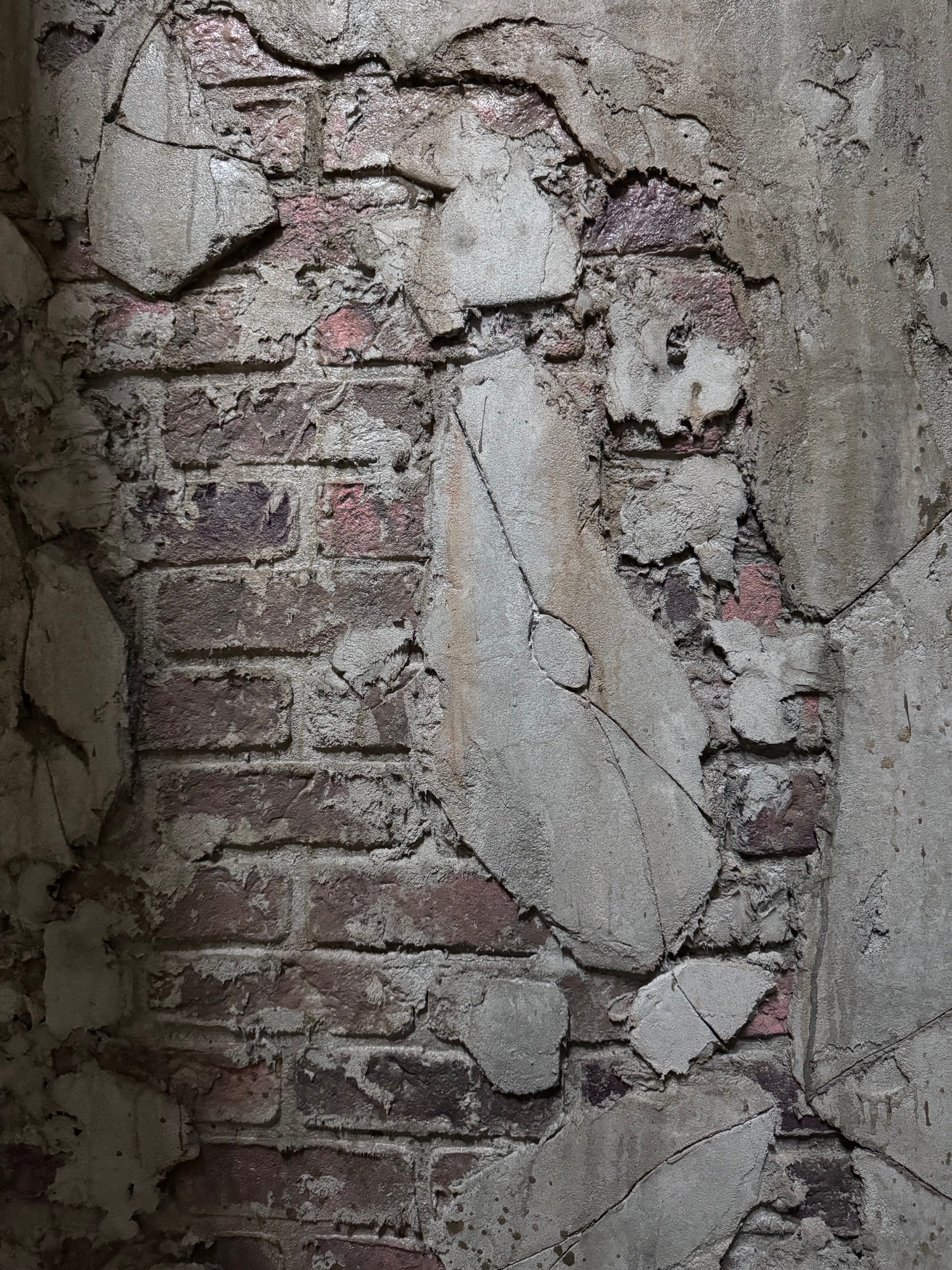 A brick wall with sections of peeling and cracked plaster, revealing the underlying bricks and some graffiti marks.