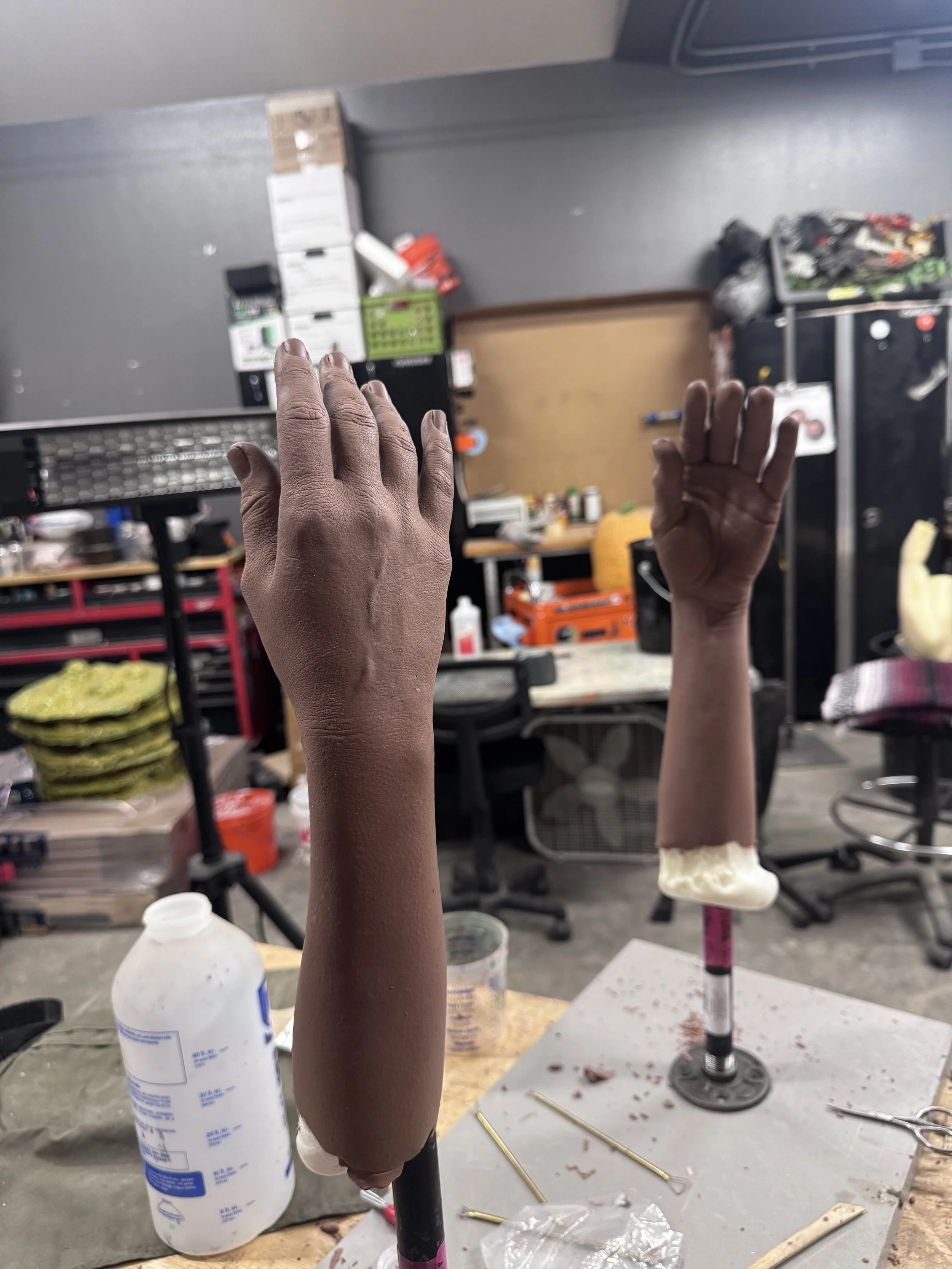 A workshop with two prosthetic arms on stands, surrounded by tools and materials.