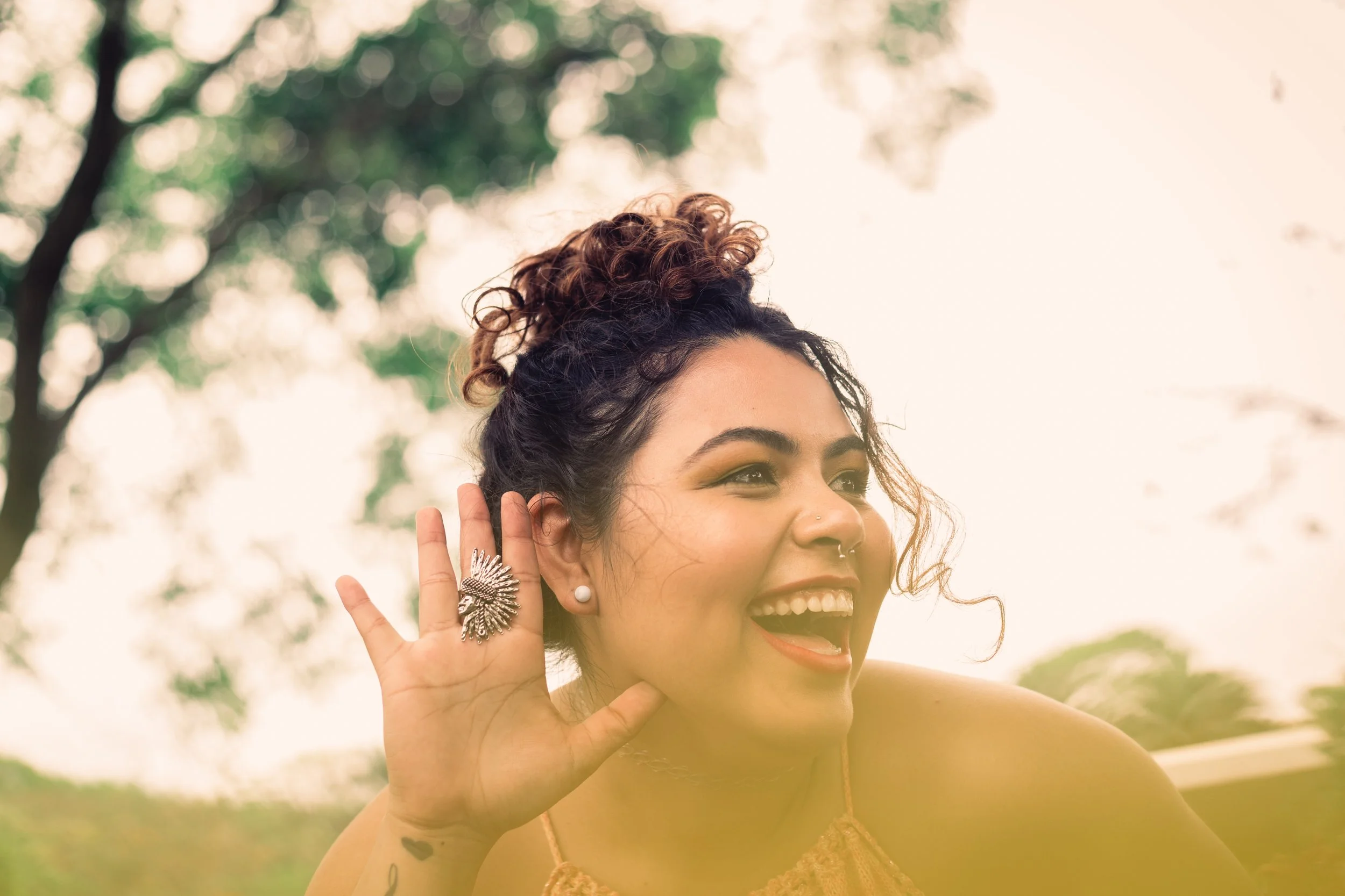 Mulher sorridente com cabelo cacheado em um coque, usando brincos de pérola, uma roupa amarela e uma peça de joalheria no dedo, segurando a orelha com a mão, ao ar livre com árvores ao fundo.