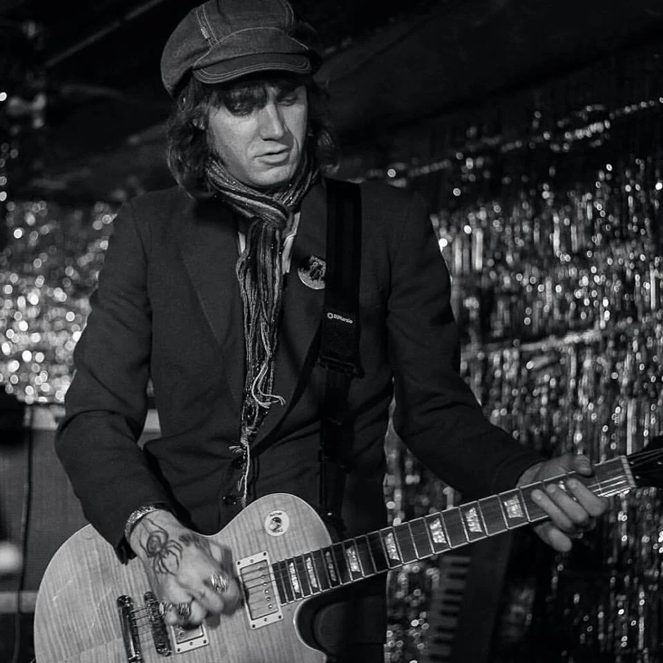 A man in a newsboy cap playing an electric guitar on stage with a shiny tinsel curtain backdrop in black and white.