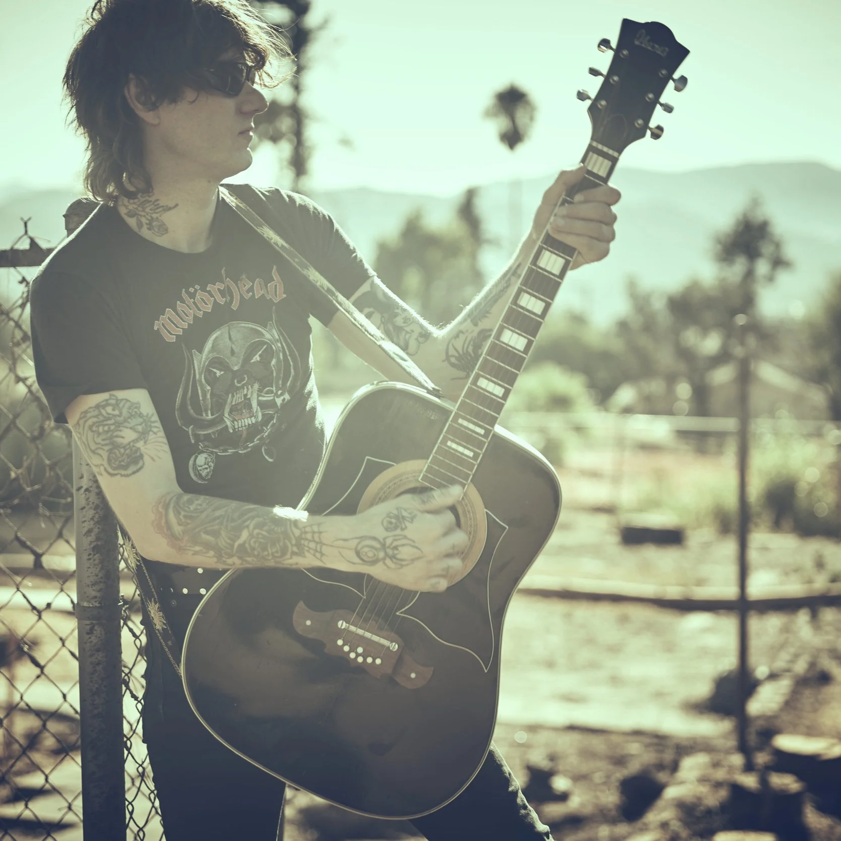 A person with tattoos and dark sunglasses playing an acoustic guitar outdoors near a chain-link fence, with trees and mountains in the background.