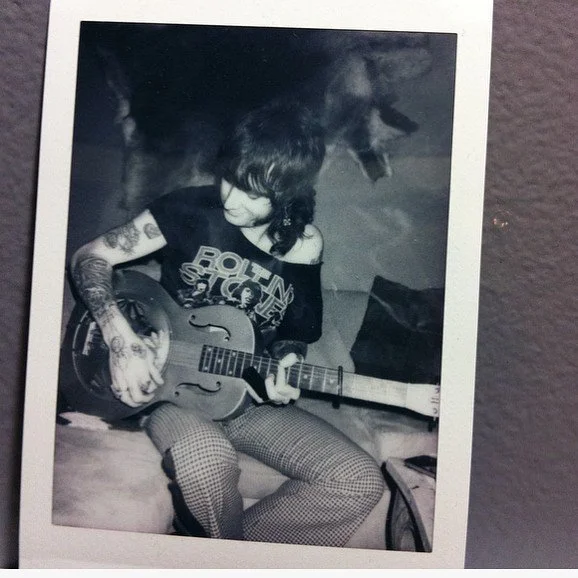 Black and white photo of a young woman sitting on a bed playing an electric guitar. She has tattoos on her arms, is wearing a T-shirt and patterned pants, and has shoulder-length hair.