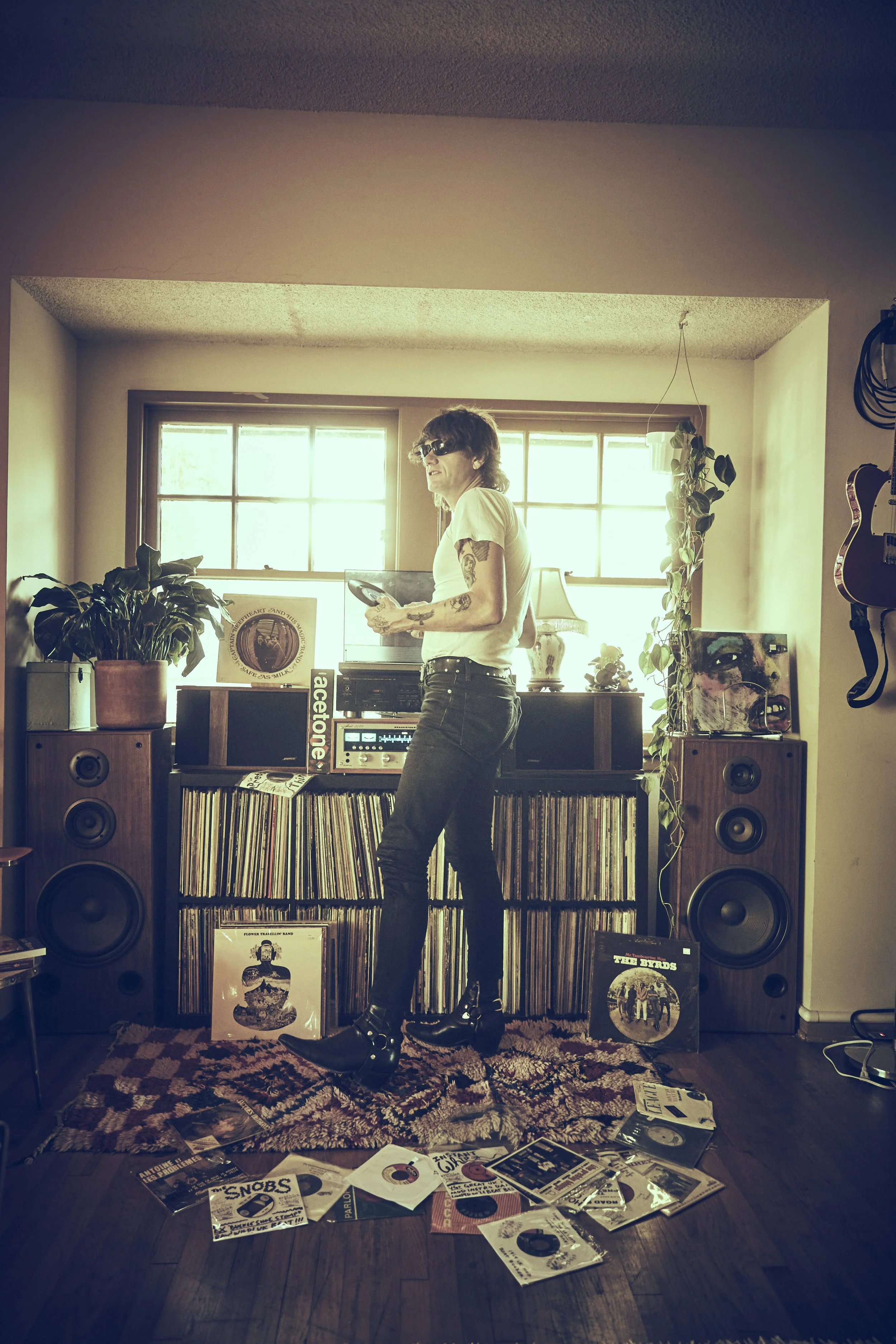A person with tattoos and sunglasses standing in a room with vinyl records, speaker systems, and musical equipment, holding a smartphone, with sunlight coming through a window behind.