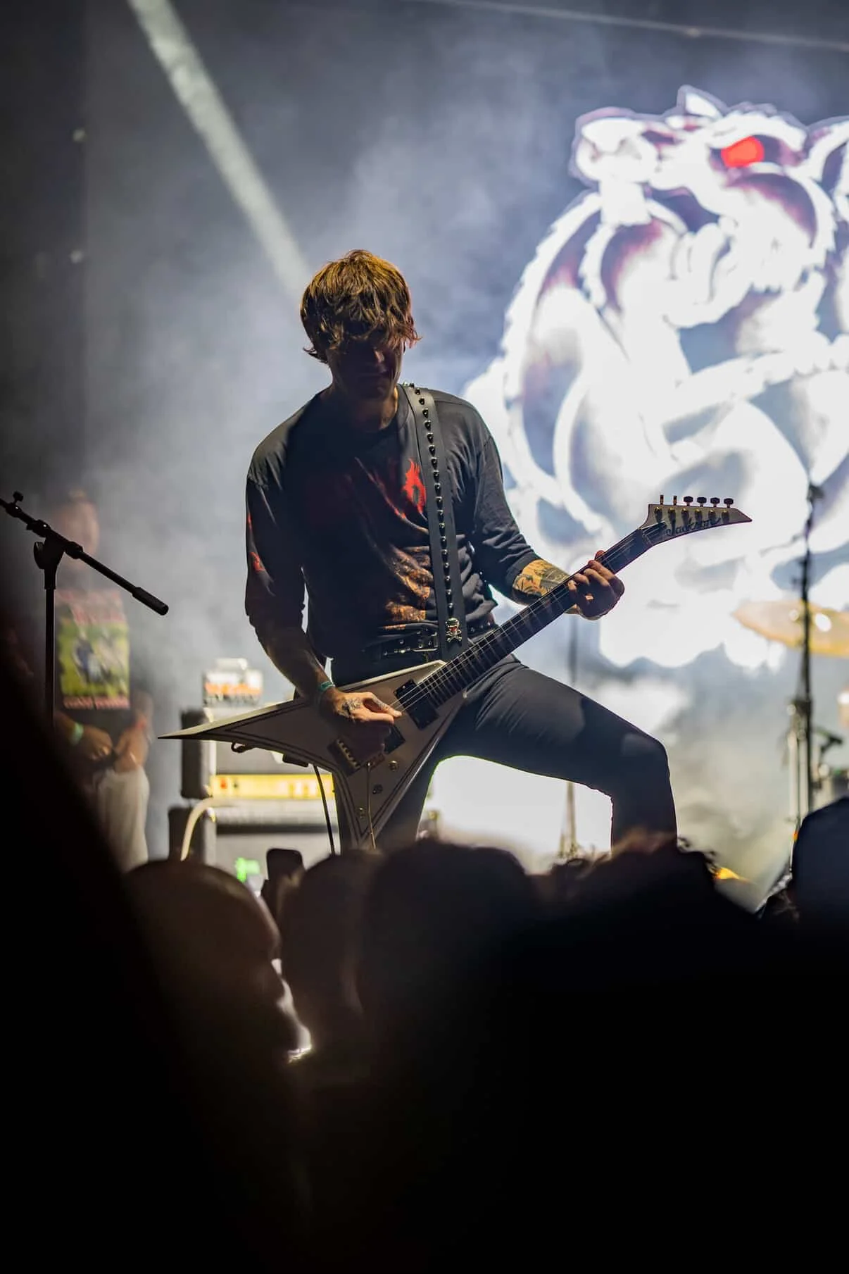 A musician playing a Flying V electric guitar on stage during a concert, with a large animated logo of a wolf in the background and concert lighting creating a dramatic effect.