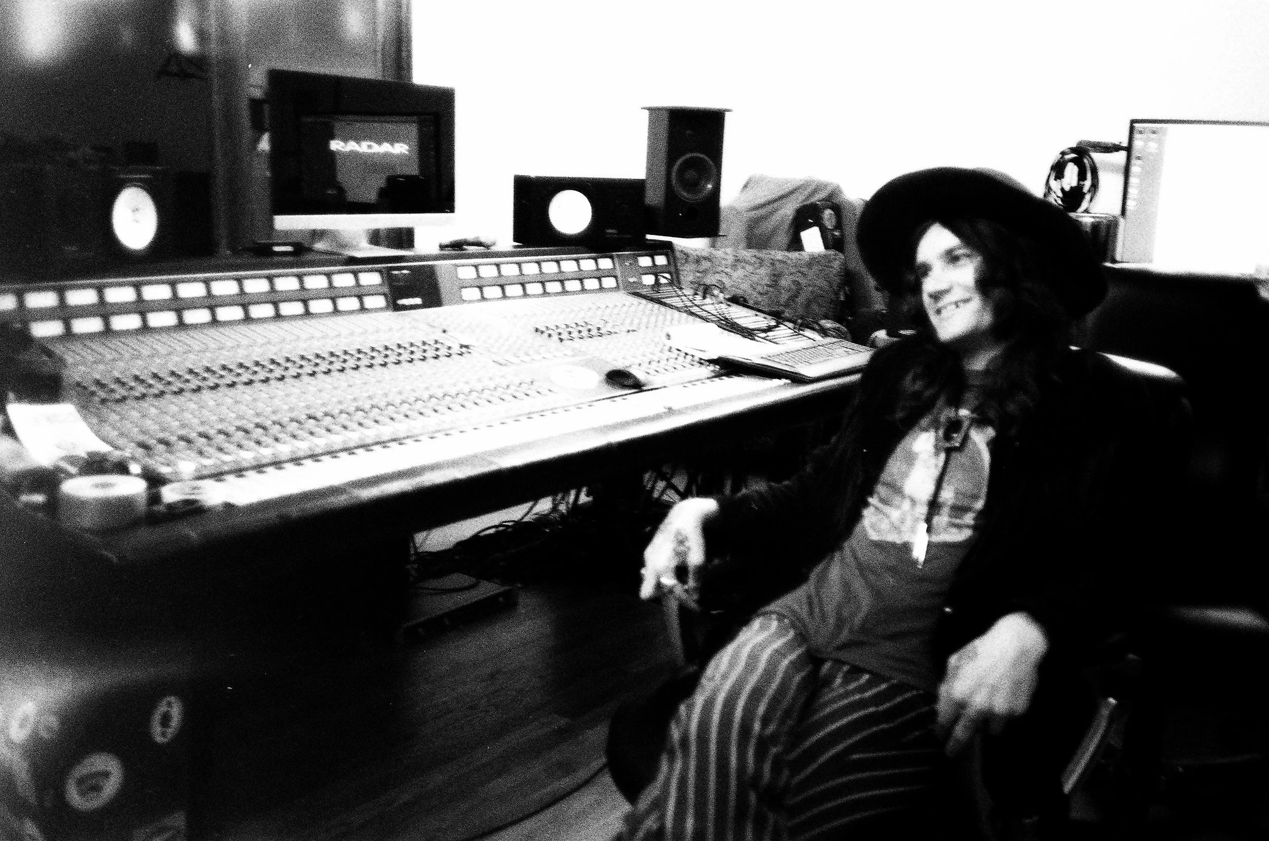 A woman sitting in a recording studio with a large audio mixing console in front of her, wearing a hat and striped pants, smiling.