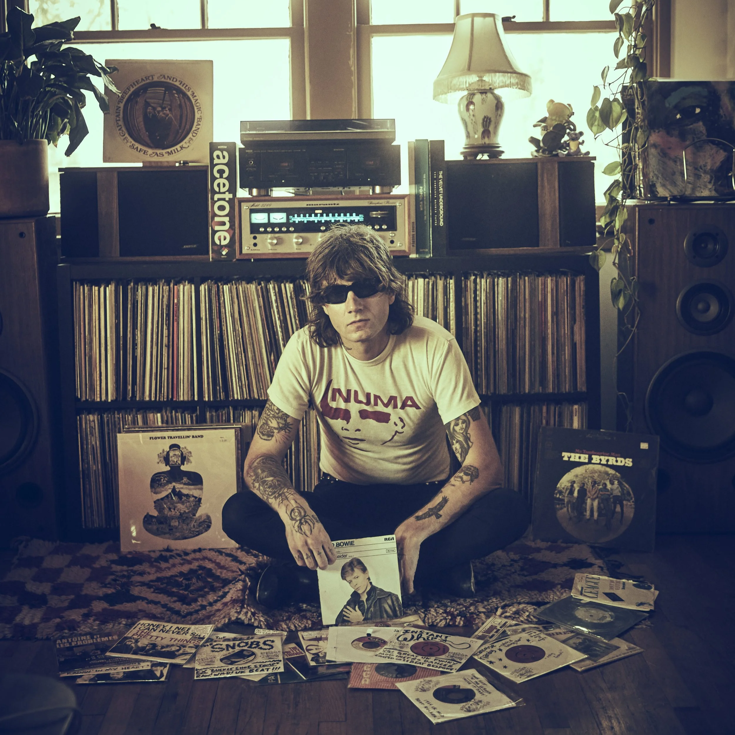 A young man with tattoos and dark sunglasses sits cross-legged on a patterned rug, surrounded by vinyl records and singles, in a room with a large vinyl collection behind him. He is holding a David Bowie album and wearing a cream-colored T-shirt with the word 'NUMA' and a face graphic. The room features vintage speakers, a stereo system on a shelf, a lamp, and potted plants near a window.