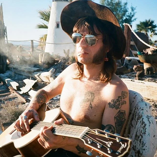 A shirtless man wearing sunglasses and a wide-brimmed hat playing an acoustic guitar outdoors. In the background, there is debris and a burned structure, suggesting a fire incident.