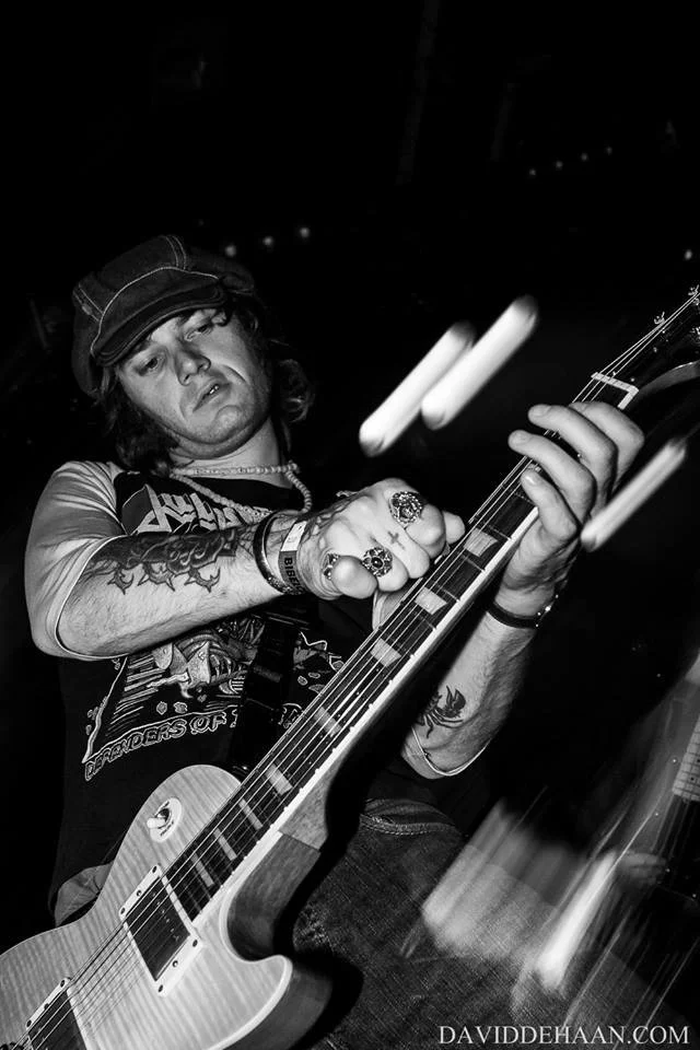 A young man with long hair wearing a cap and jewelry playing an electric guitar on stage in a low-light setting.