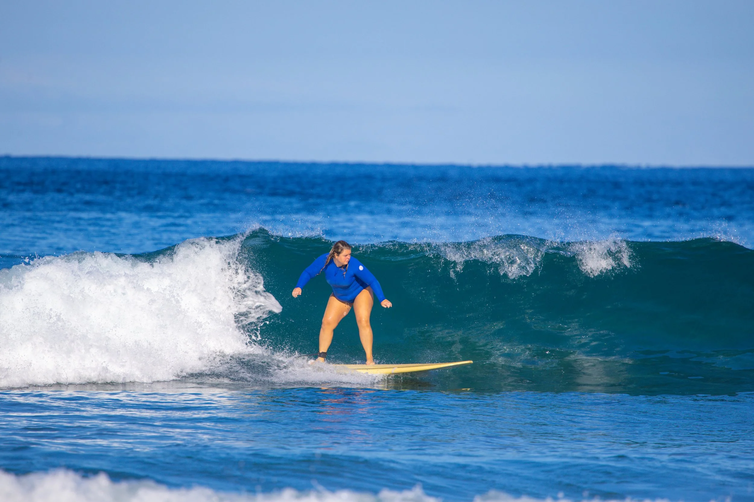 A woman wearing a blue jacket surfing on a yellow board on the ocean waves under a blue sky.