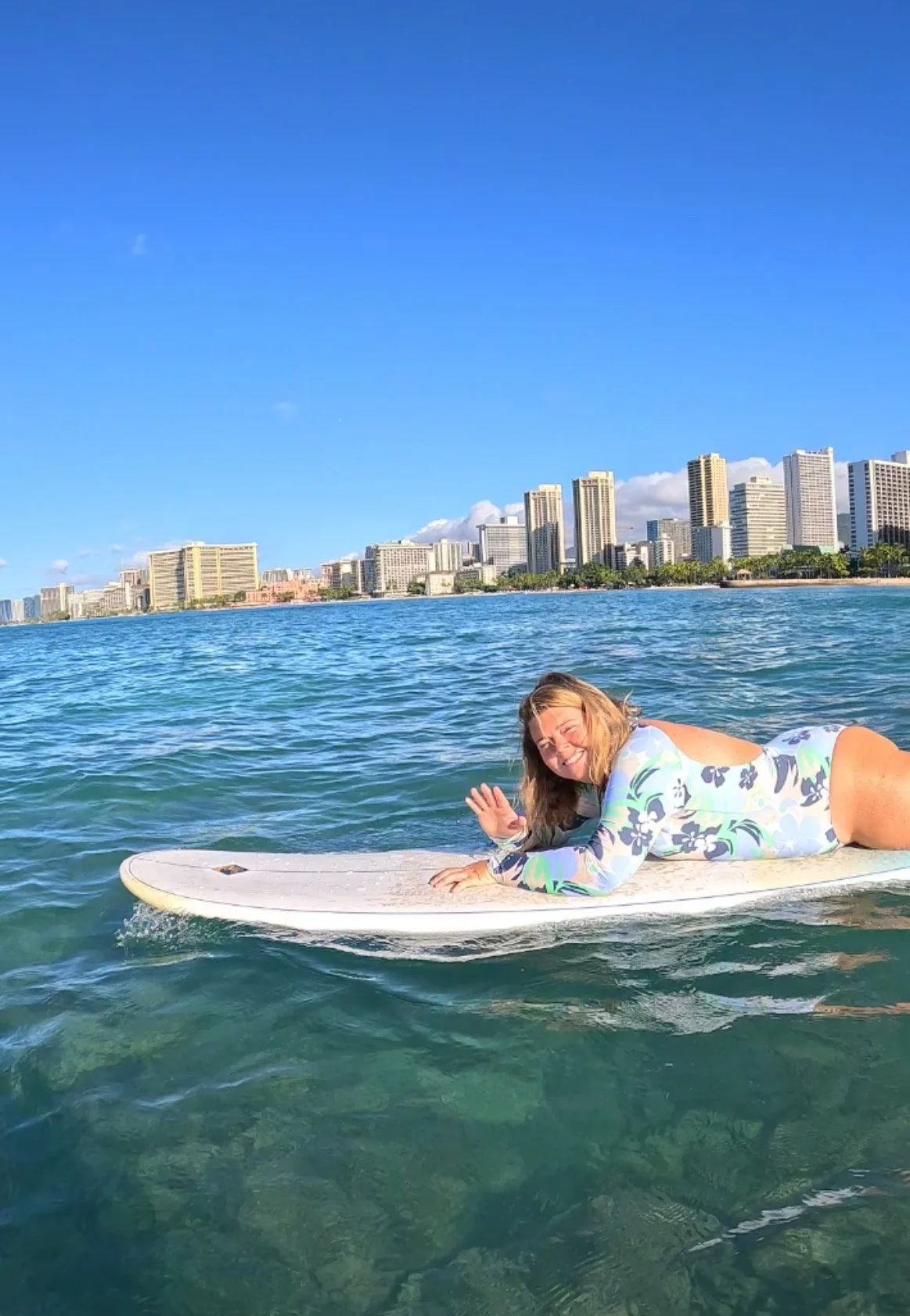 A woman in a floral wetsuit lying on a surfboard in the ocean with a city skyline in the background, smiling and waving.