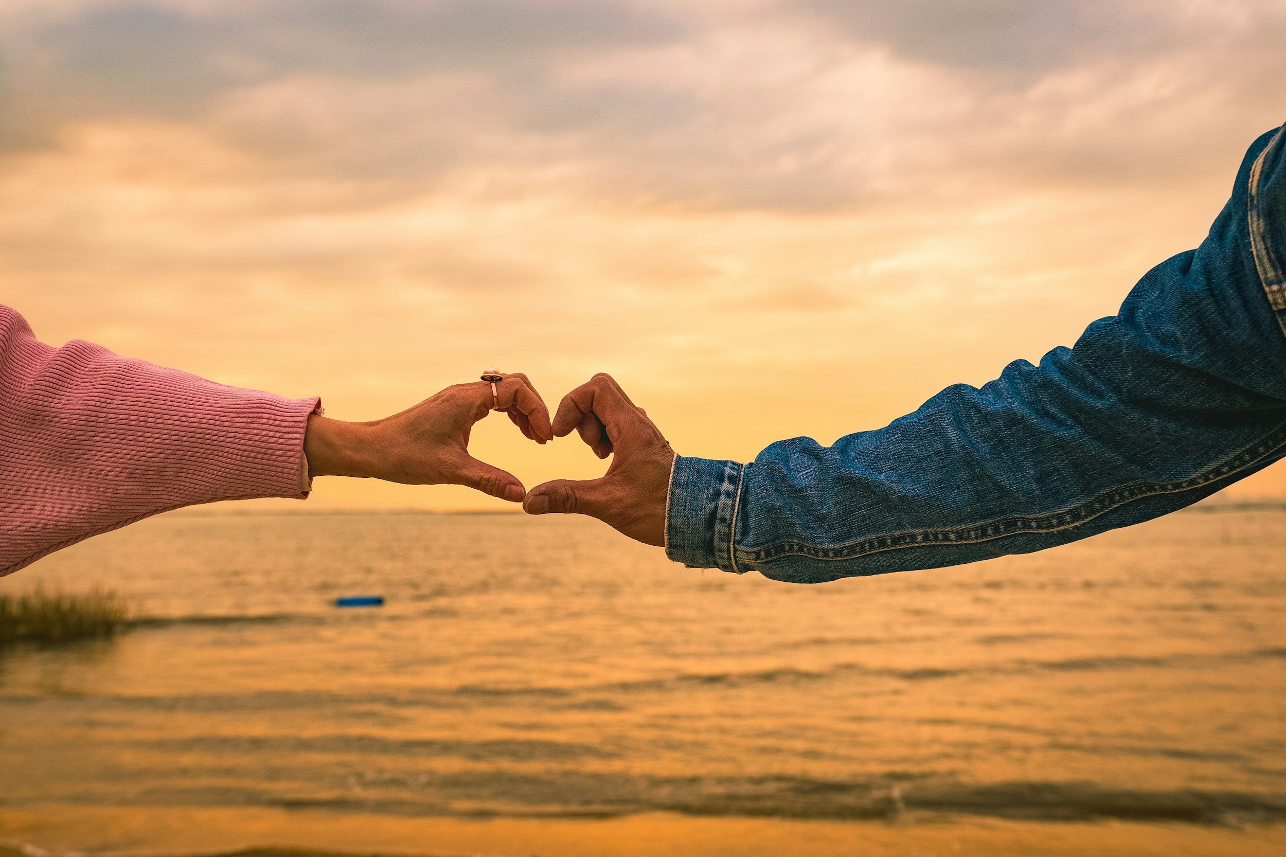 Two people forming a heart shape with their hands, one wearing a pink sweater and the other in a denim jacket, near a beach at sunset.