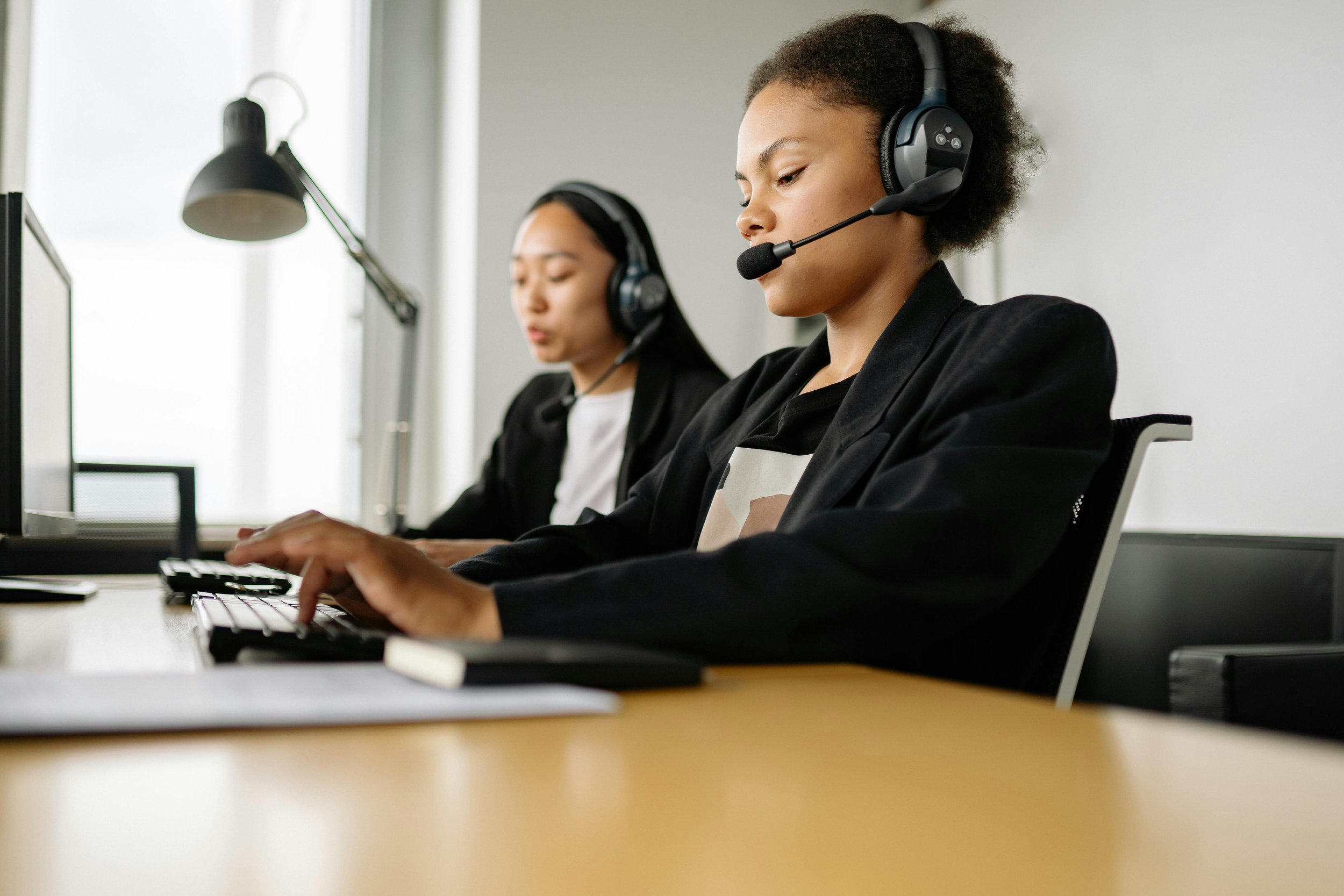 Two women wearing headsets working at computers in an office setting.