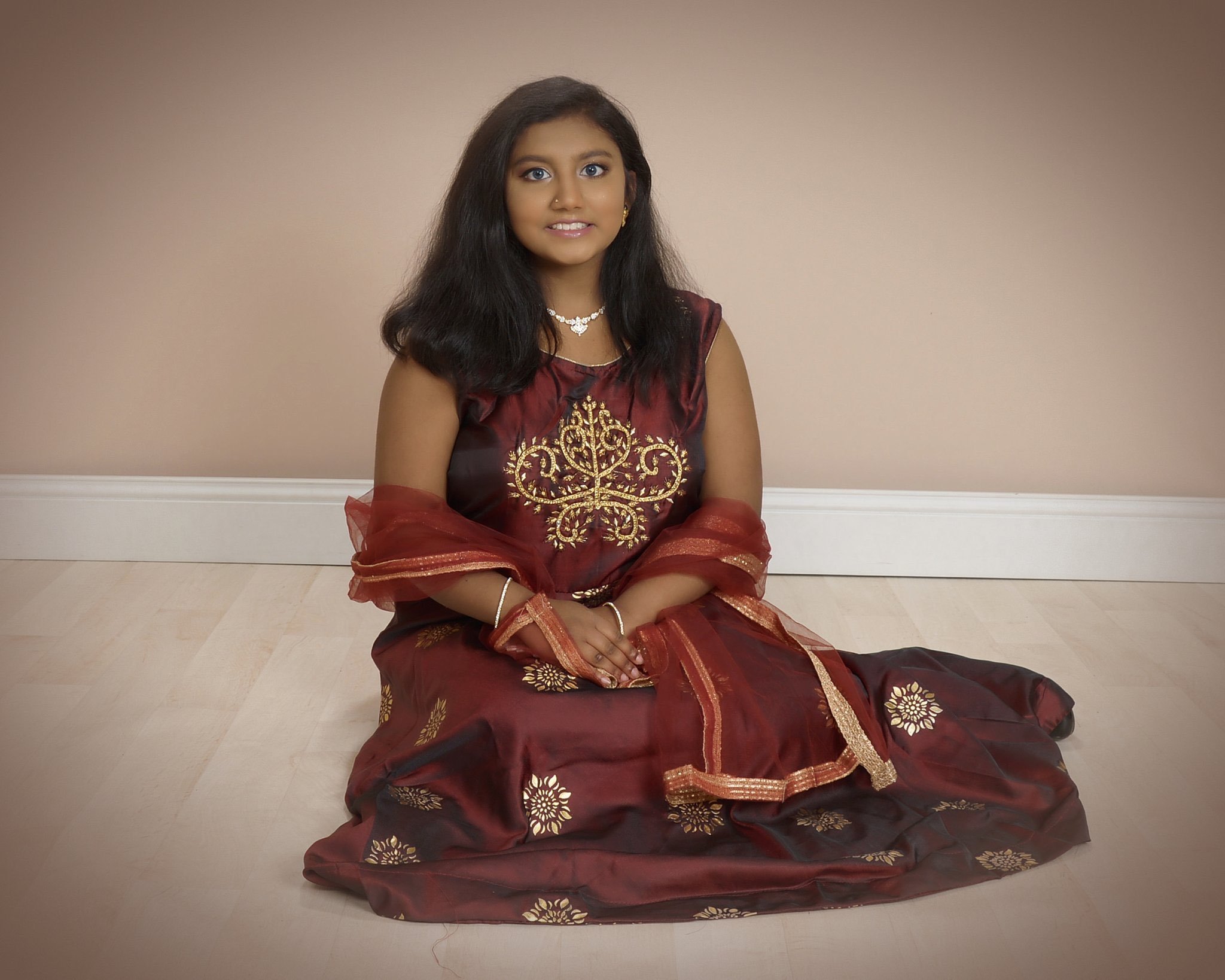 A young girl sitting cross-legged on a wooden floor against a beige wall, wearing a traditional maroon and gold dress, with jewelry and a sheer shawl.