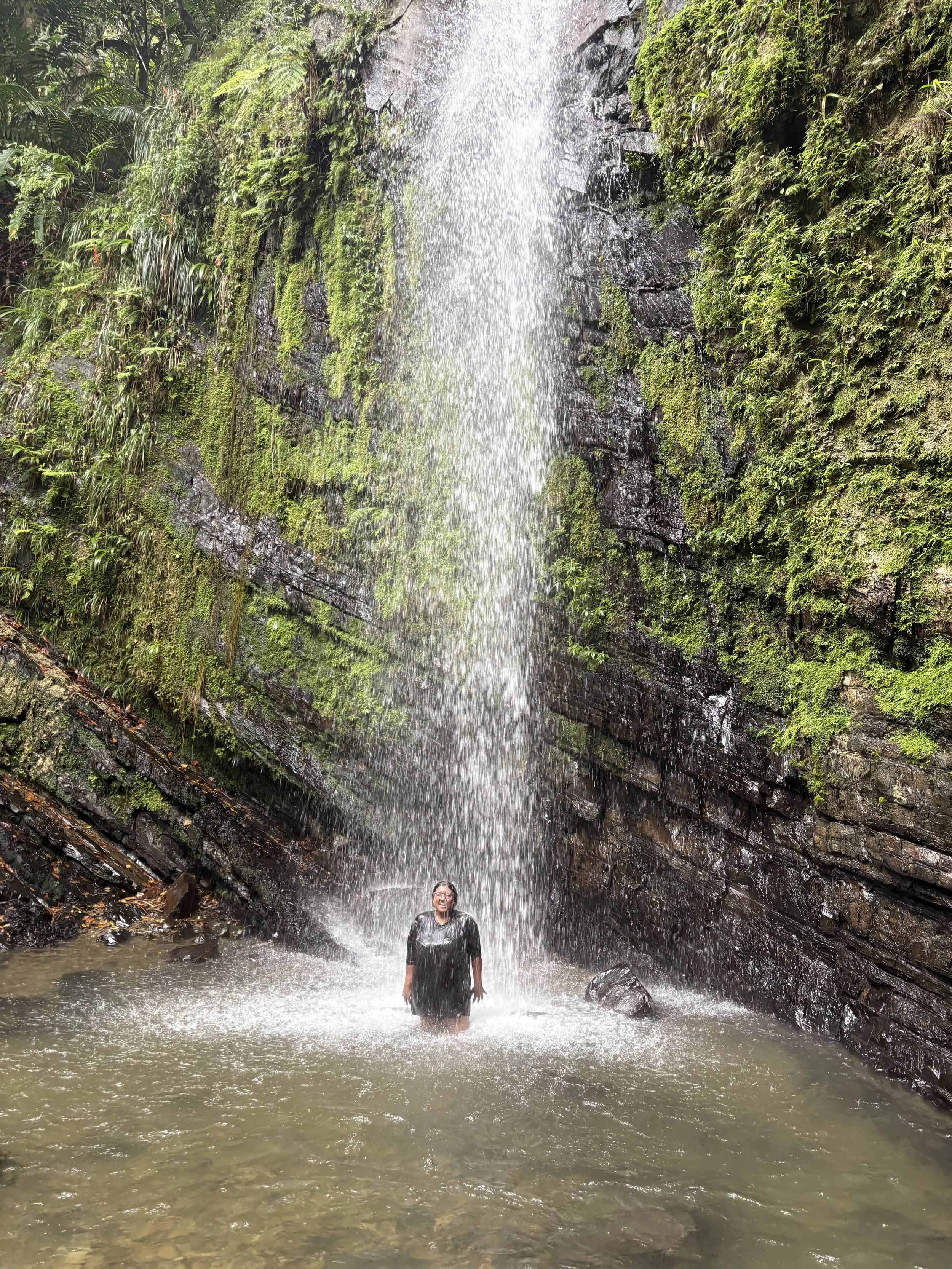 Person standing beneath a waterfall in a rocky and mossy canyon.