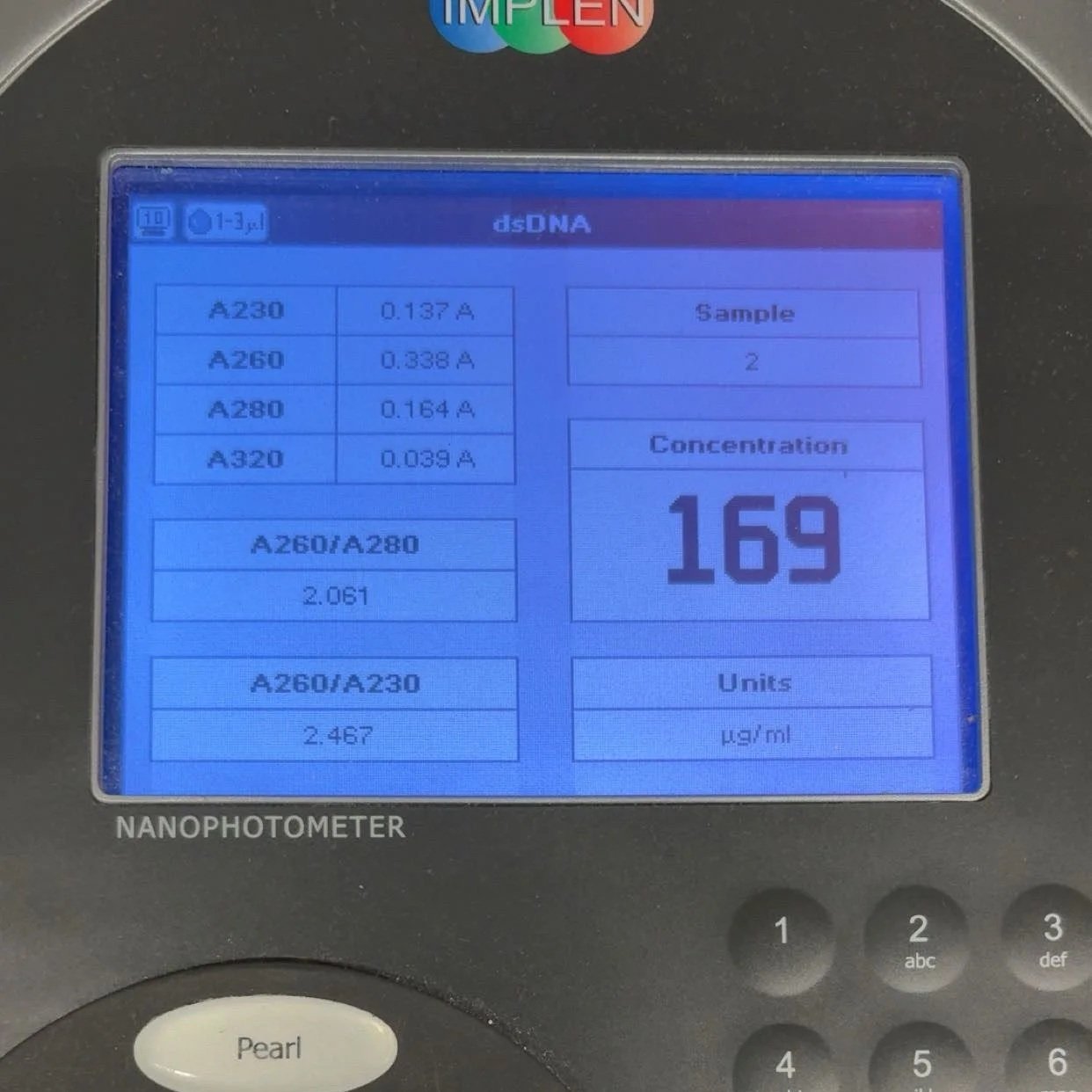 Close-up of a nanophotometer display screen showing DNA sample measurements, including concentrations of different DNA types and ratio calculations, with a numeric keypad and a branded logo at the top.