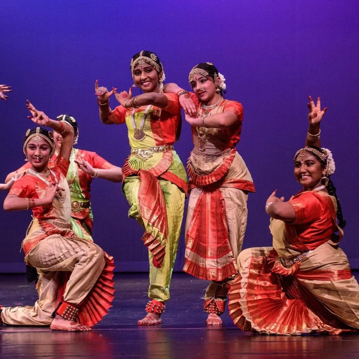 Five young girls in traditional Indian dance costumes performing on stage, with a purple background.