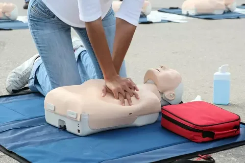 A person practicing CPR on a medical training mannequin outdoors, with a red emergency bag and a bottle of hand sanitizer nearby.