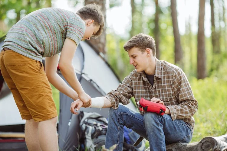 Two young men camping outdoors, one seated with a bandaged arm in a sling and holding a first aid kit, the other leaning over to support his injured arm.