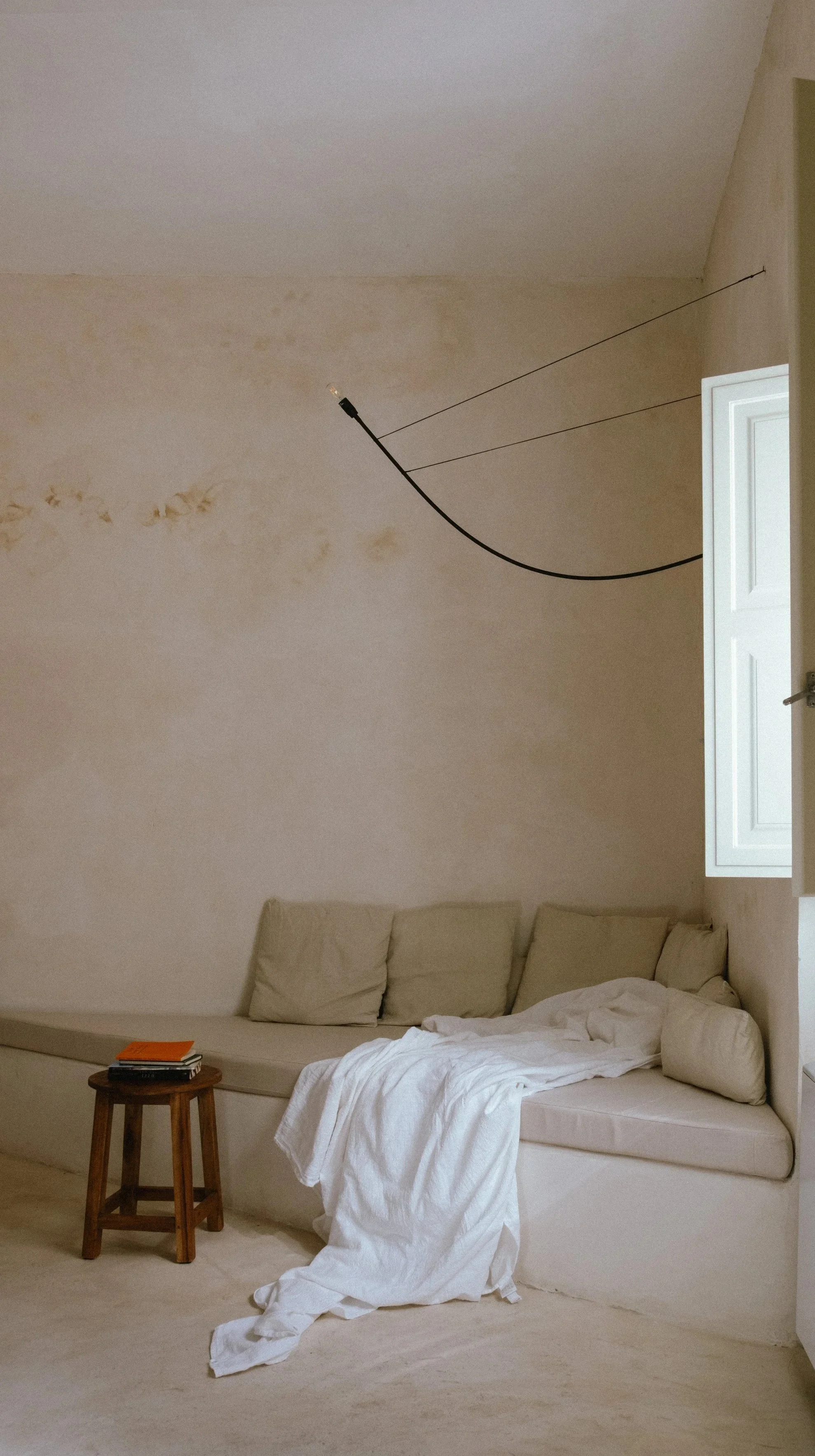 A beige room with a built-in couch, beige cushions, white bedding draped over the side, a small wooden stool with books and an orange notebook, and an open window with white shutters. A black cable hangs from the ceiling.