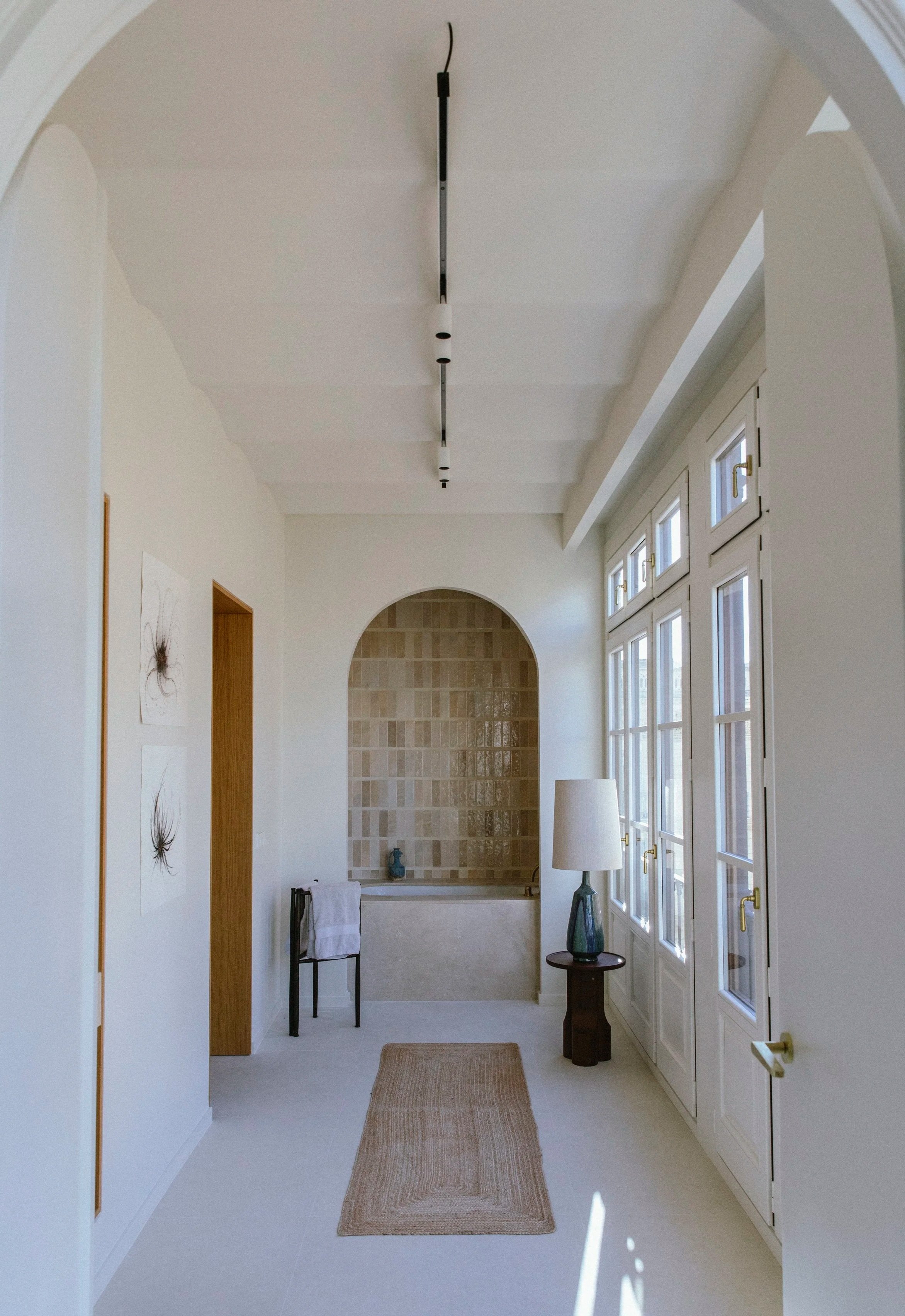 Bright hallway with large windows, a beige runner rug, a corner bathtub, a floor lamp, and decorative art on the wall.