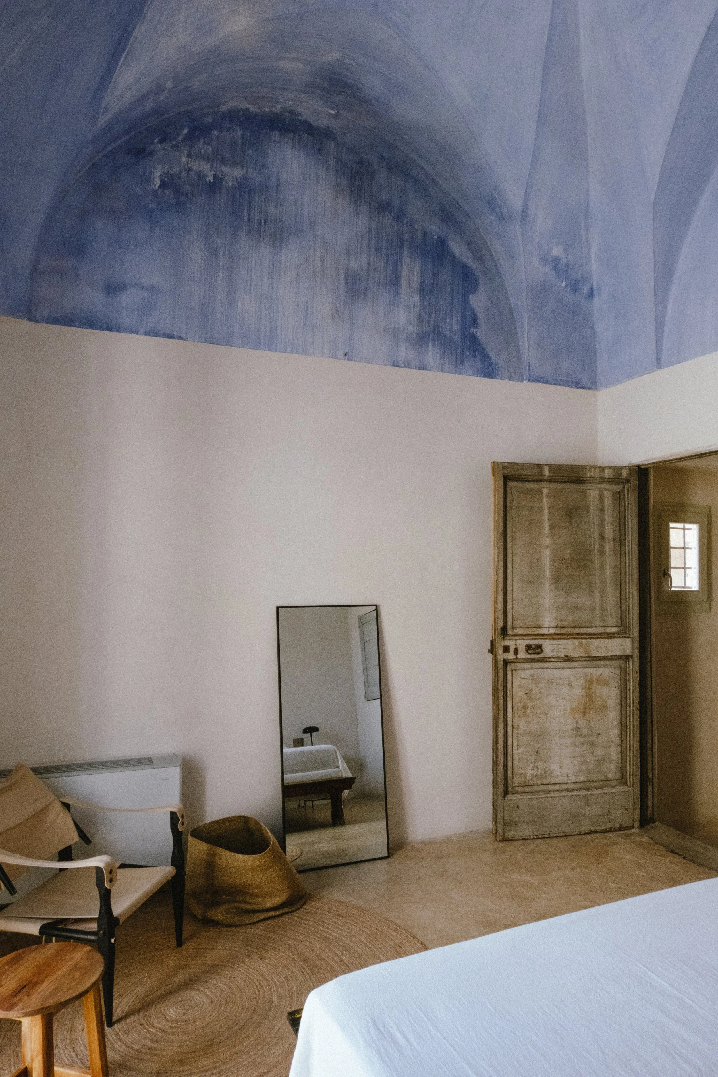 Interior of a bedroom with a white bed, a standing mirror, a wicker basket, and a wooden chair, with an aged wooden door and a small window, and a blue painted vaulted ceiling.